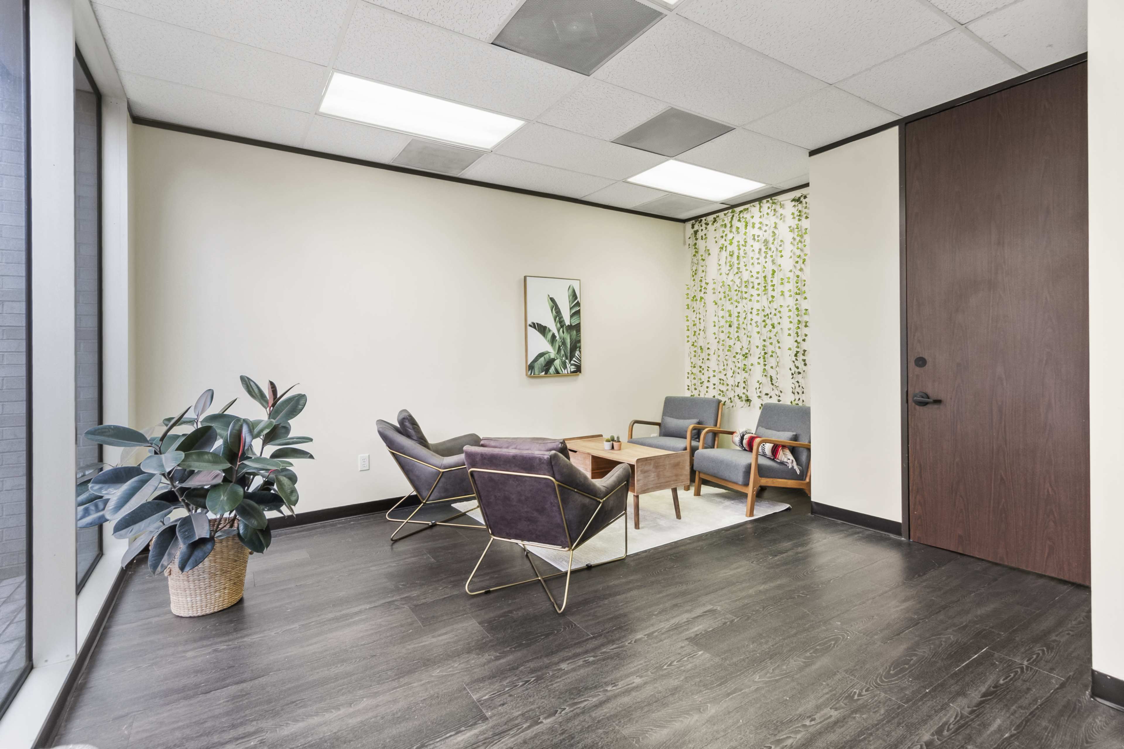 A modern waiting area features two gray armchairs, a green potted plant, and a decorative artwork of leaves on the wall.