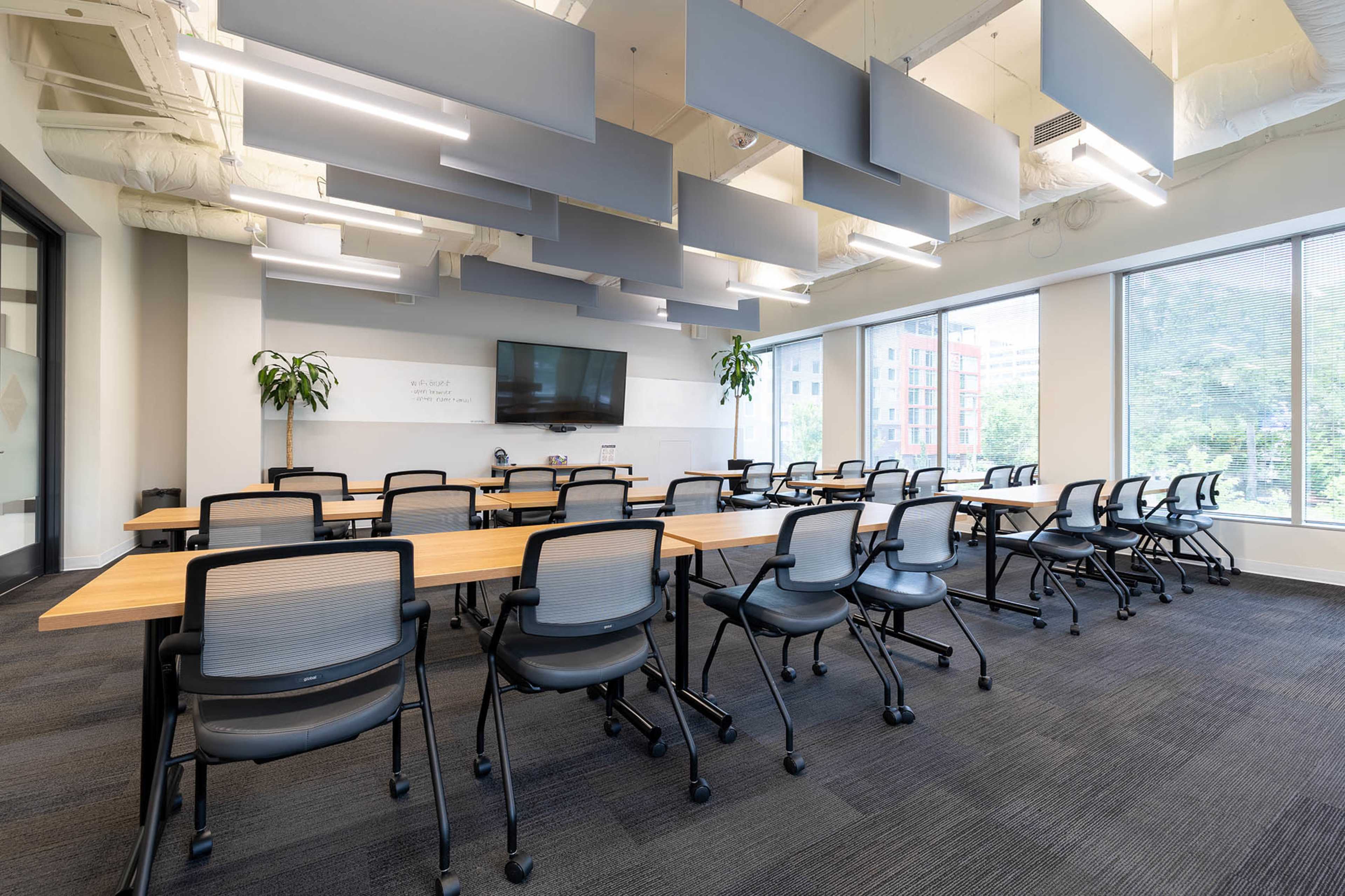 A modern conference room features a long table surrounded by ergonomic chairs, with large windows offering a view of the outdoors.