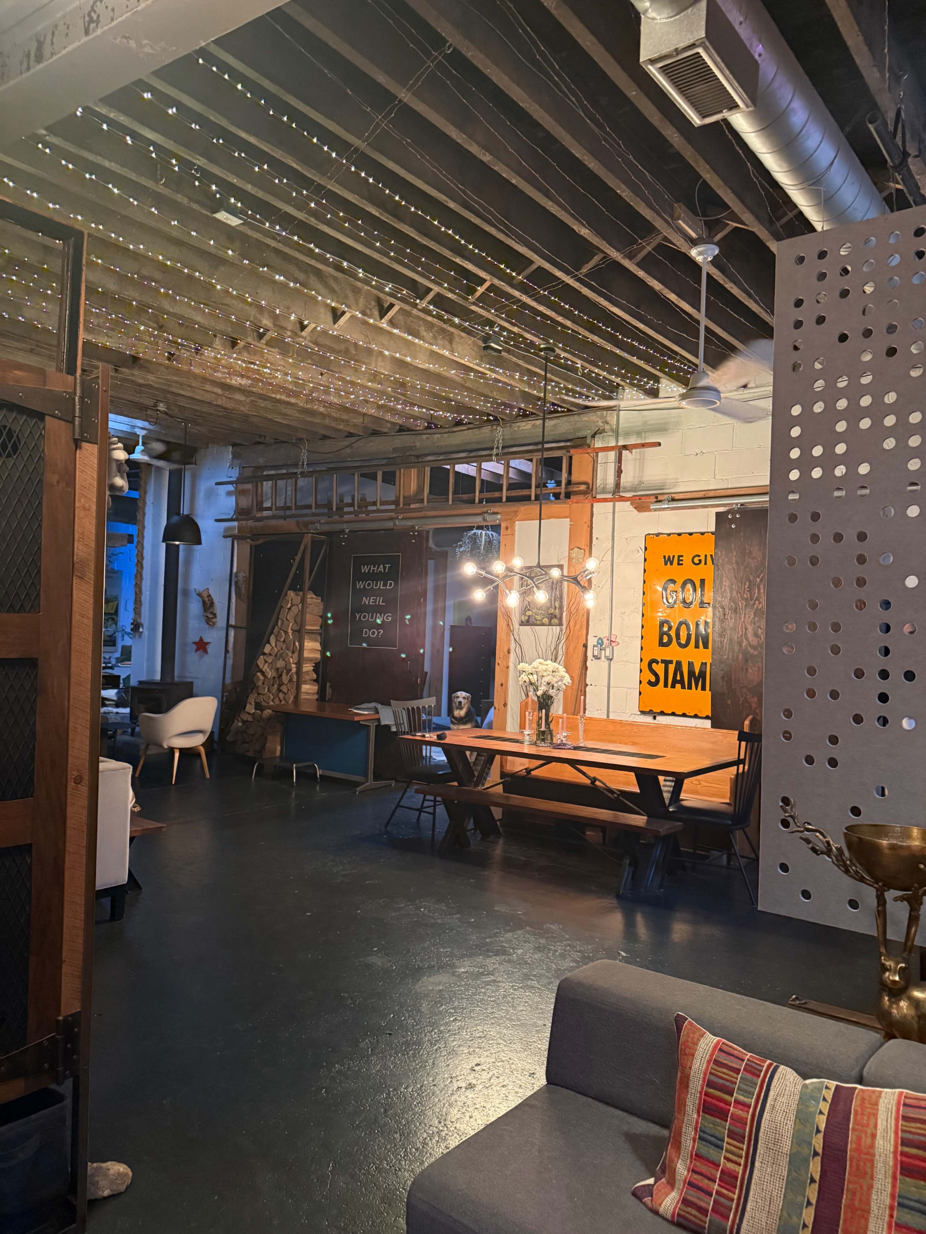 The image shows a modern, eclectic interior space with exposed beams, bright decorative lighting, and a large wooden table surrounded by chairs.