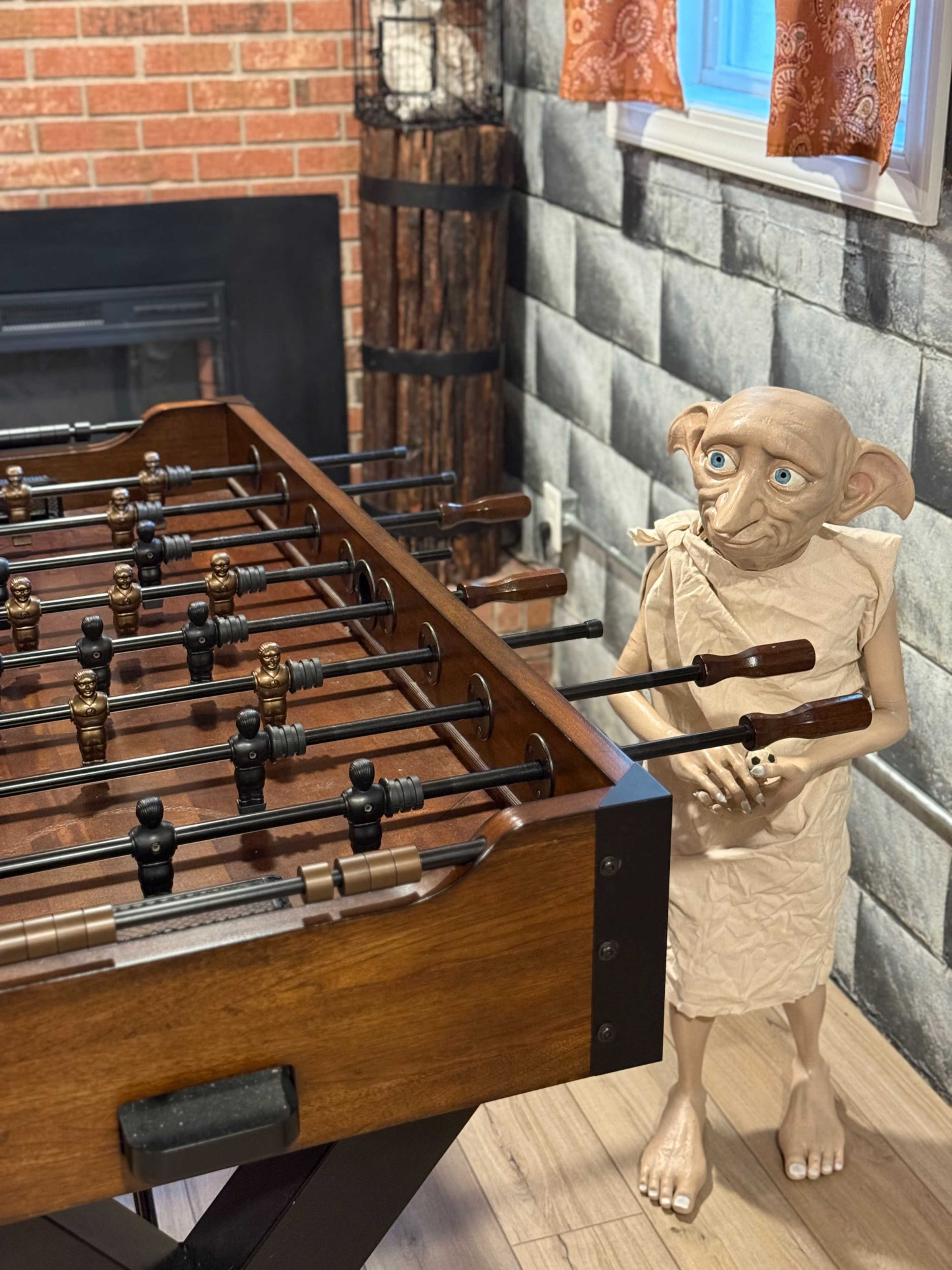 A foosball table stands in a room with brick and stone walls, beside a life-sized figure resembling a fictional house-elf.