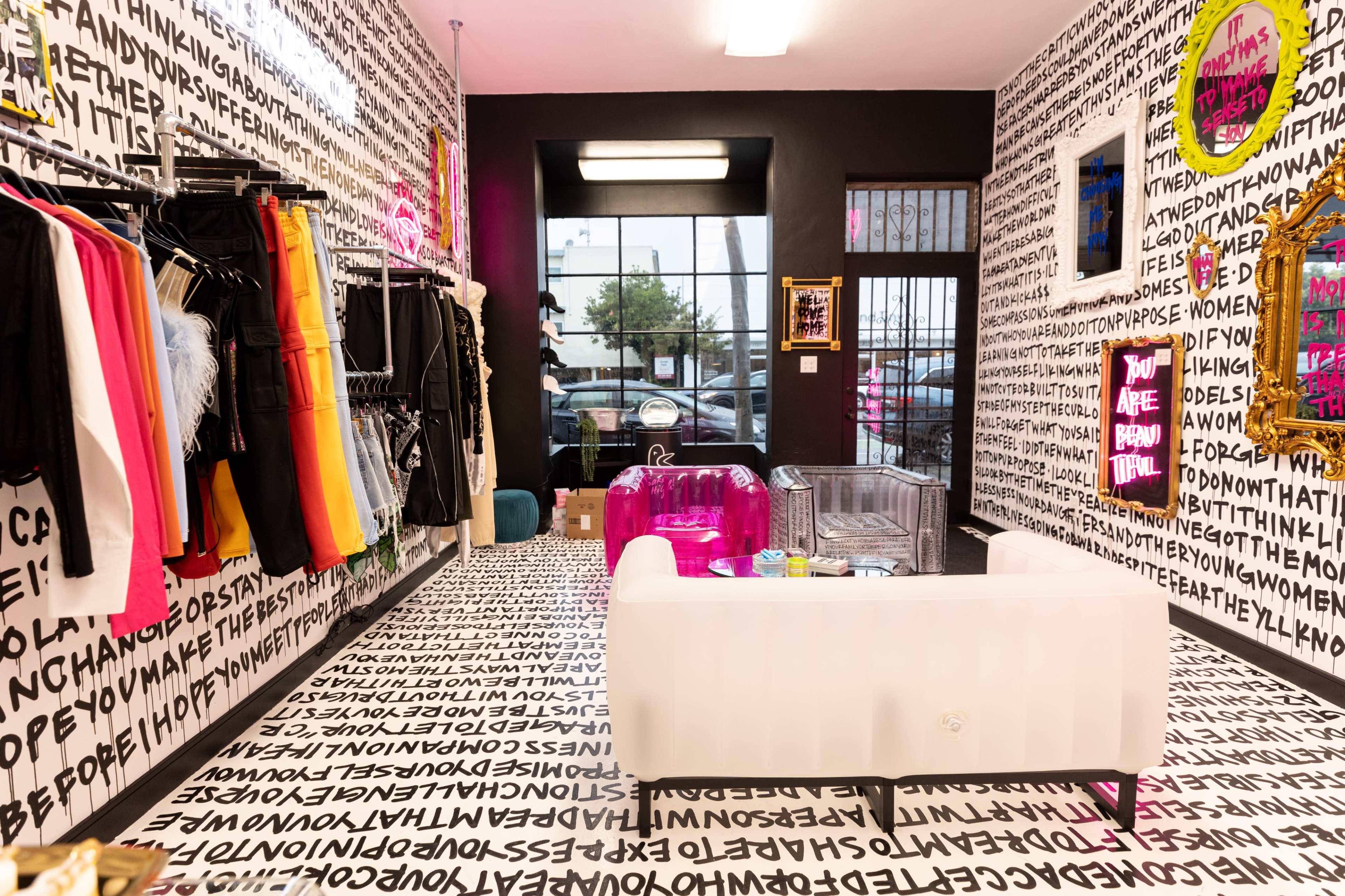A boutique interior with walls covered in black and white text, displaying colorful clothing racks on one side and a transparent seating area on the other.