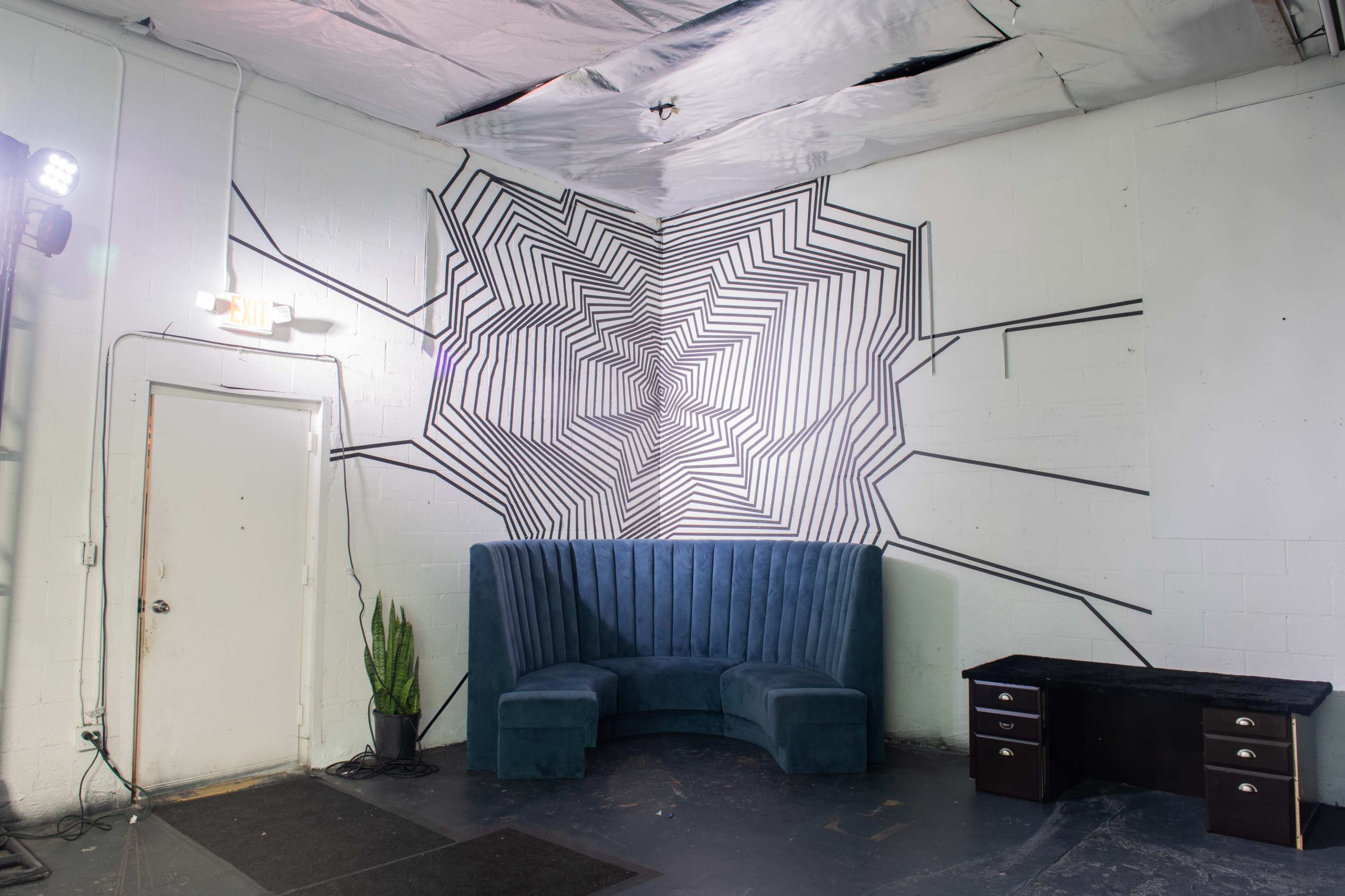 A corner of a room featuring a large geometric black and white mural on the wall, a green velvet couch, and a black table with drawers.