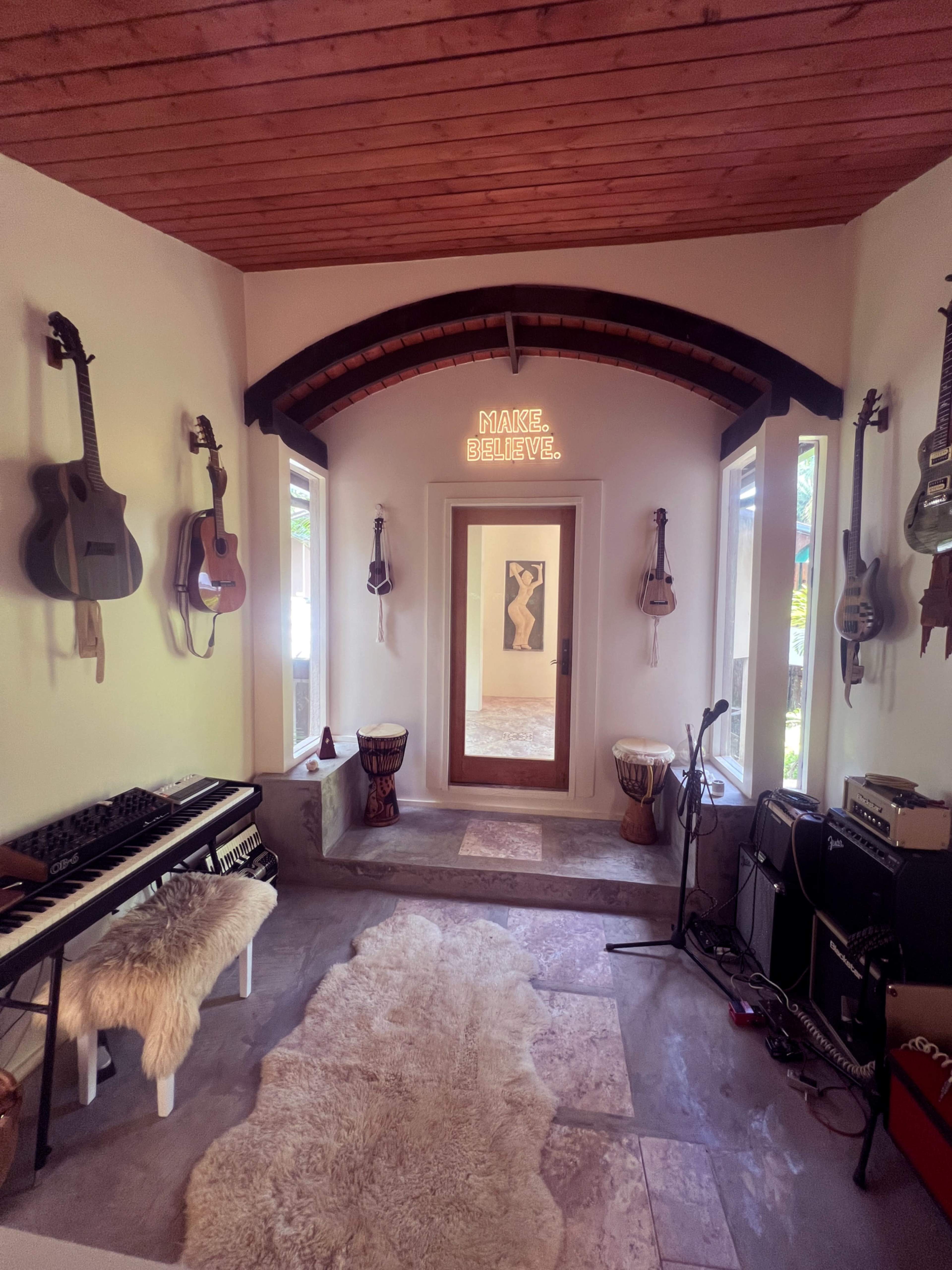 The image shows a music room featuring various guitars mounted on the walls, a keyboard on a stand, and drum instruments, with a decorative mirror at the far end and a neon sign that reads "MAKE. BELIEVE."