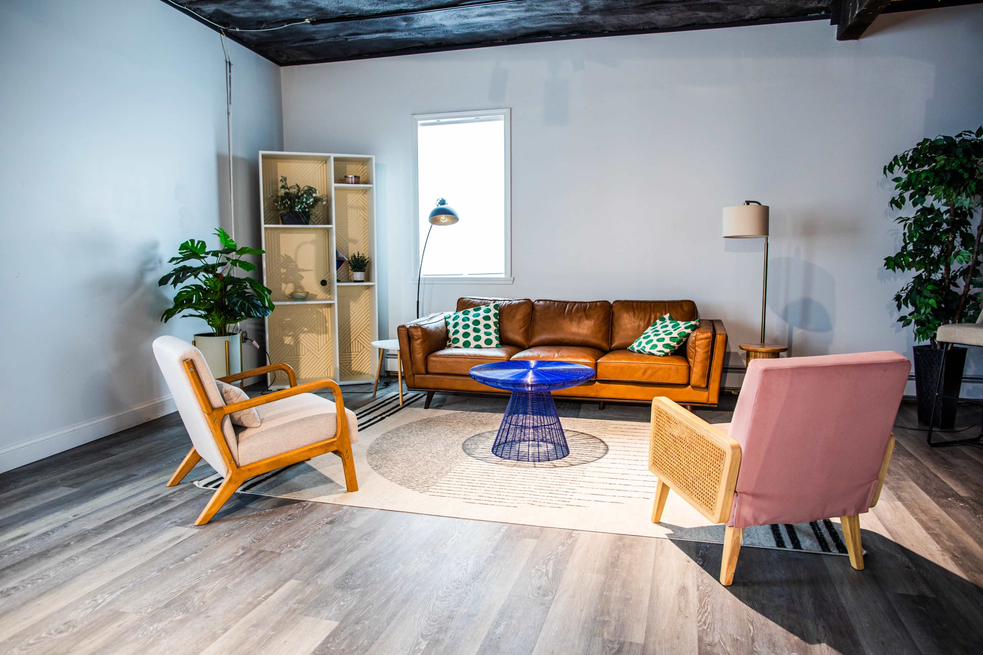 The image shows a modern living room with a brown leather sofa, two accent chairs, a blue coffee table, a patterned rug, and a tall plant beside a window.