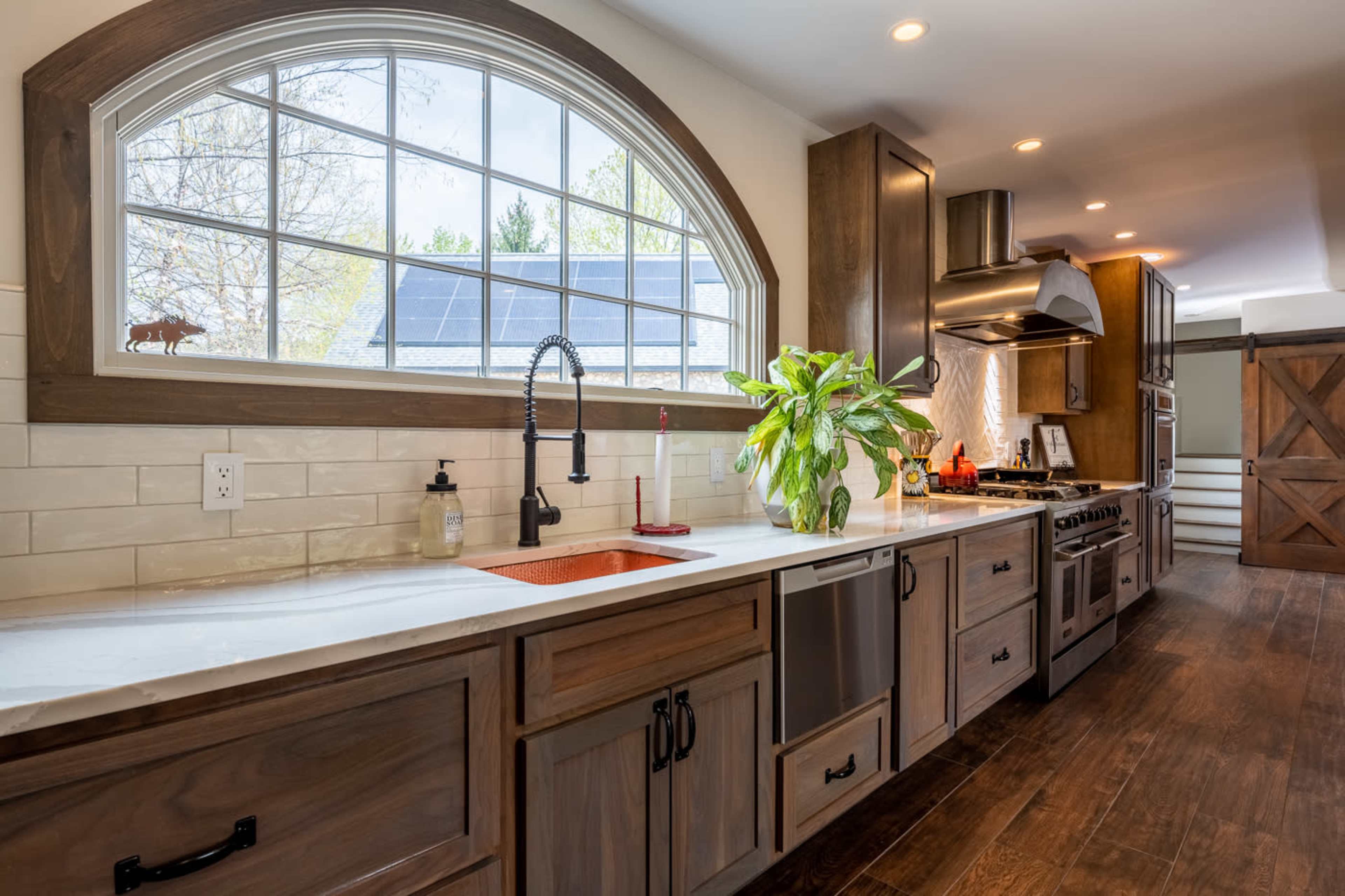 A modern kitchen features wooden cabinetry, a large window with an arched top, a farmhouse sink with a copper basin, and a dark hardwood floor.