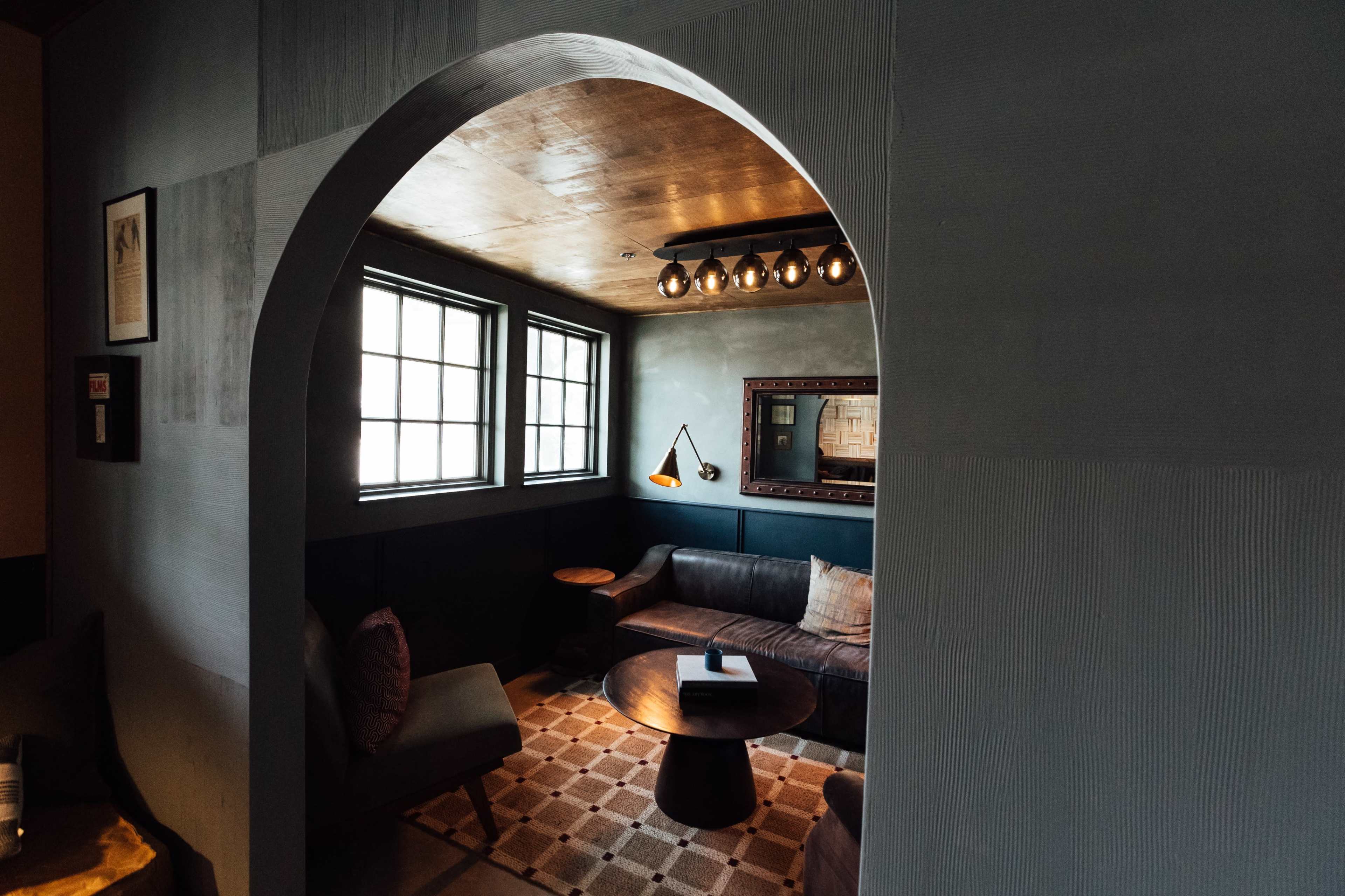 A cozy seating area is framed by an arch, featuring a sofa, wooden tables, and large windows.