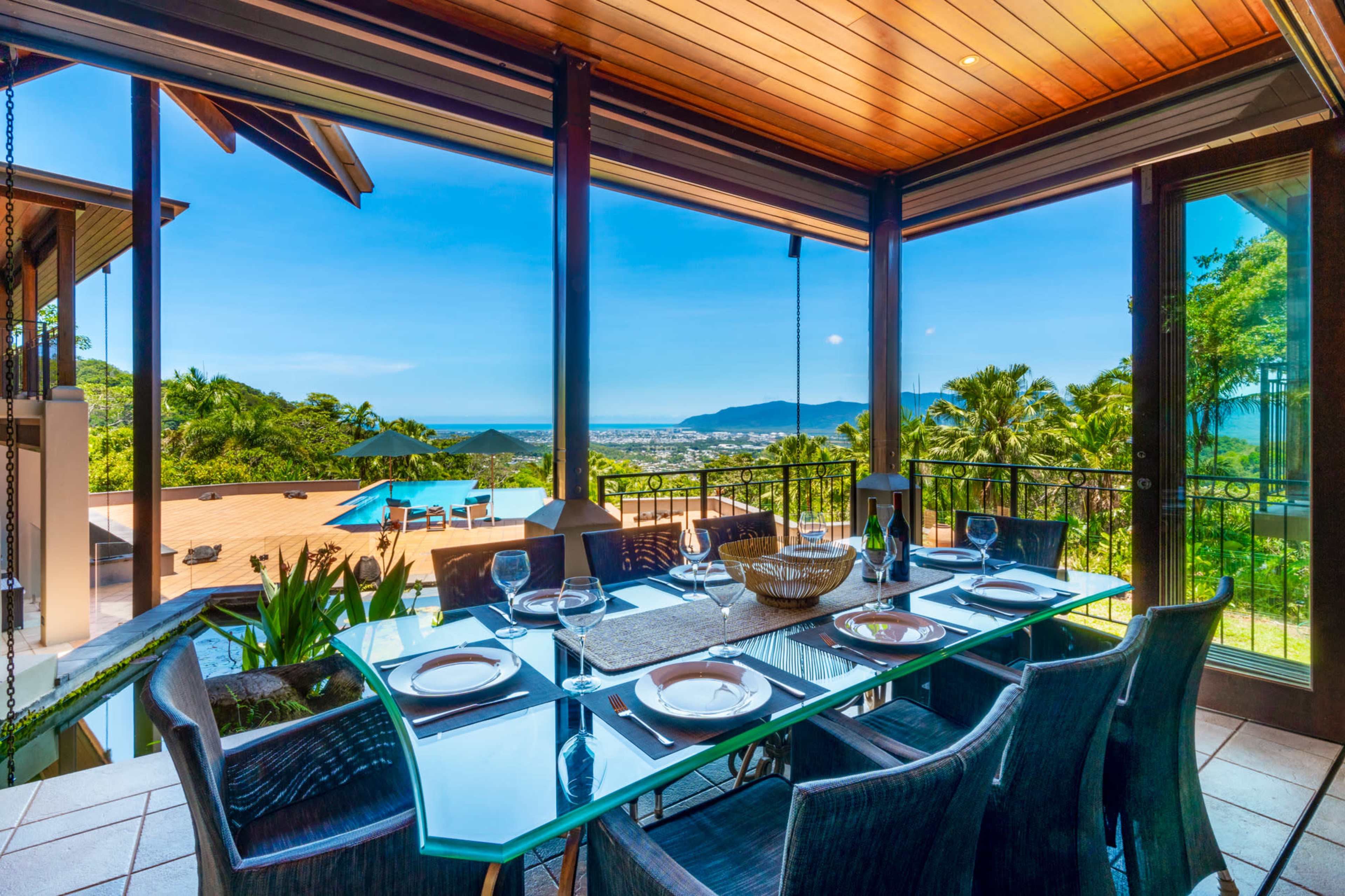 A spacious dining area features a glass table set for dinner, overlooking a scenic view of the mountains and ocean.