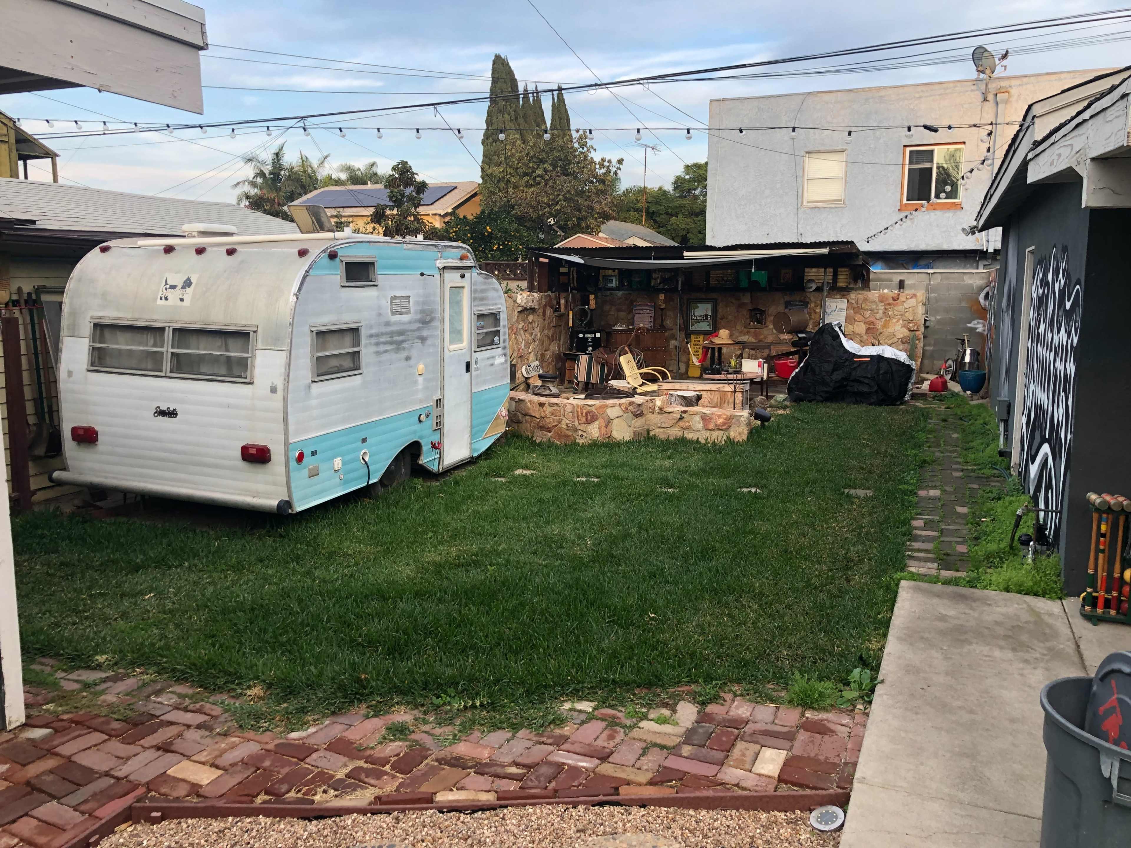 A vintage blue-and-white trailer sits on a grassy yard next to a stone area with outdoor furniture and a covered workspace.