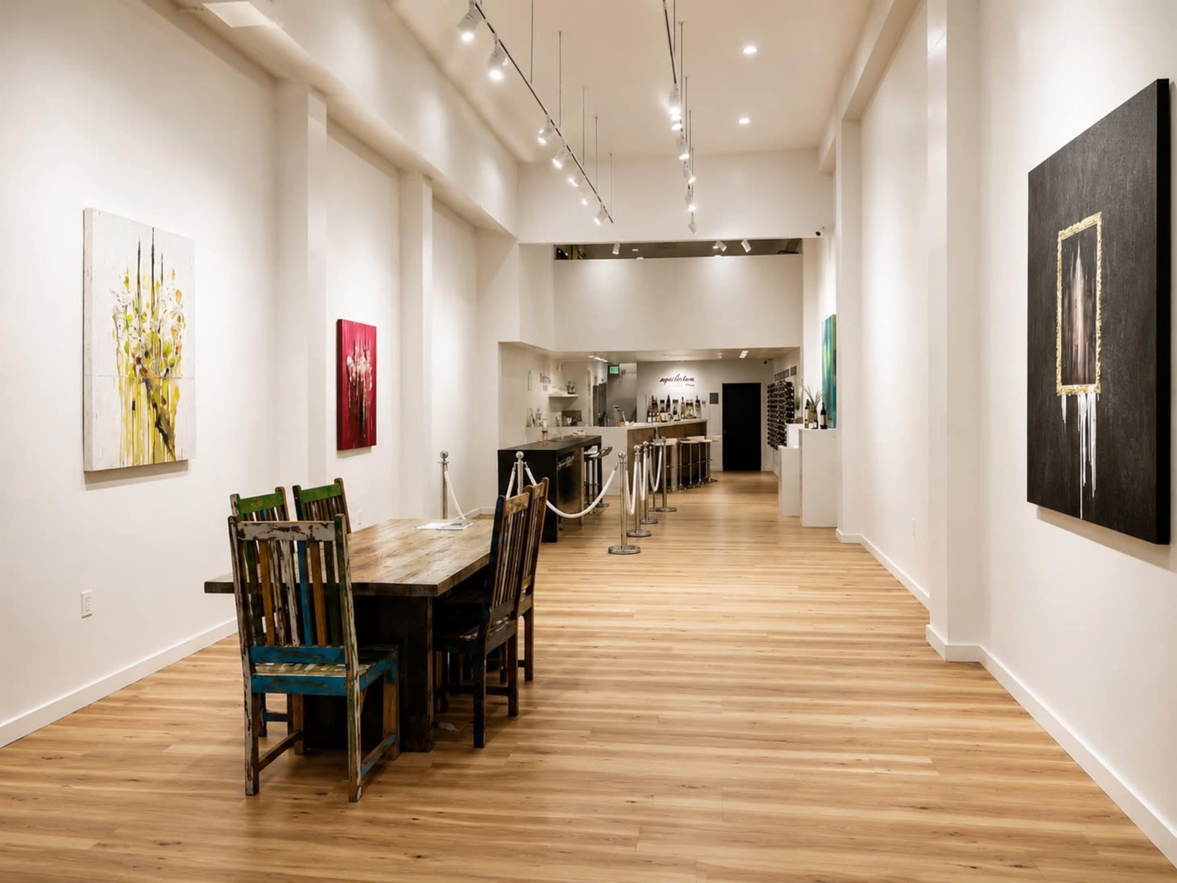 The image shows a spacious gallery with wooden floors, featuring colorful paintings on the walls and a long wooden table with mismatched chairs in the foreground.