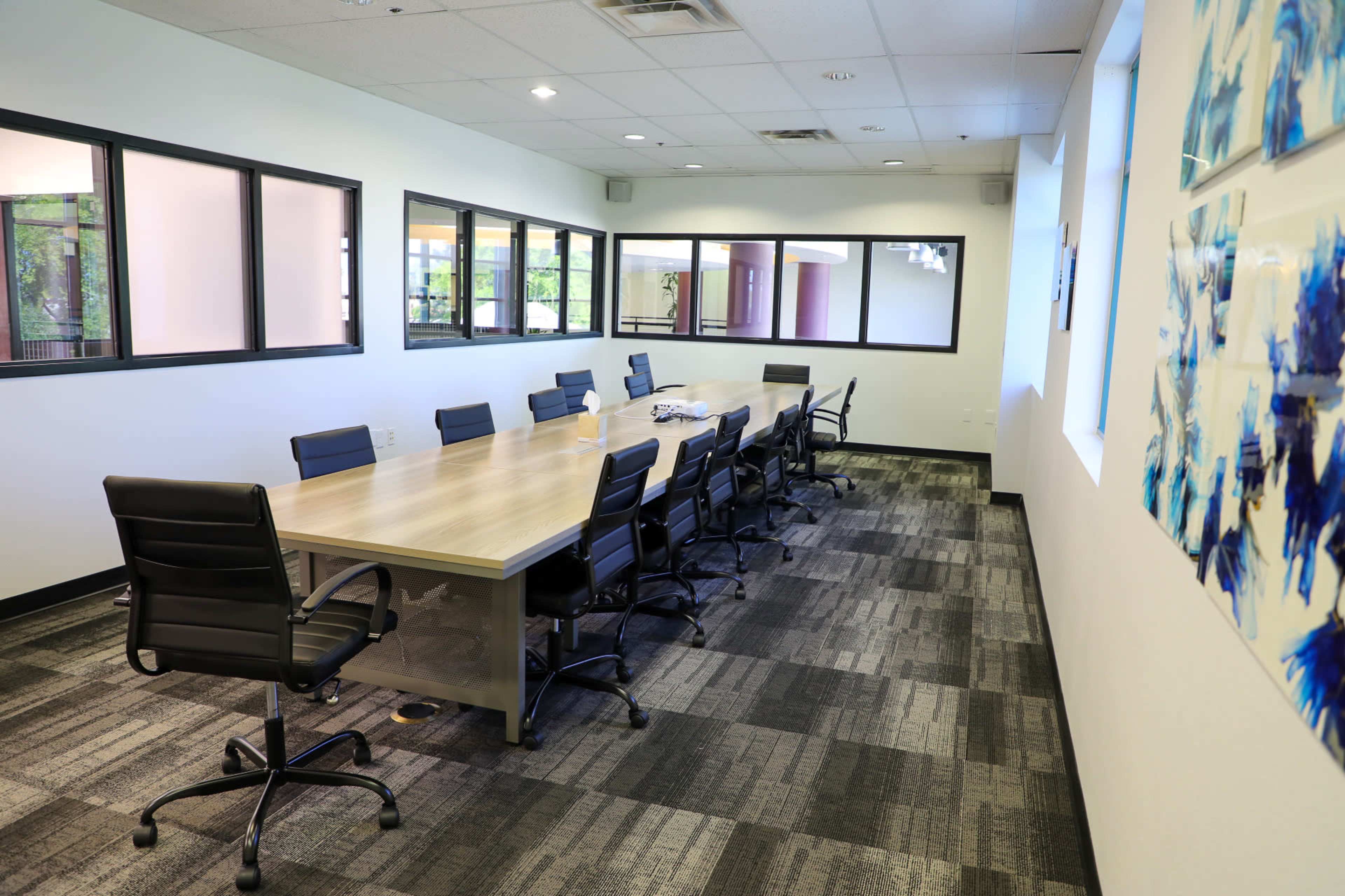 A spacious conference room with a long wooden table surrounded by black chairs and large windows along the walls.