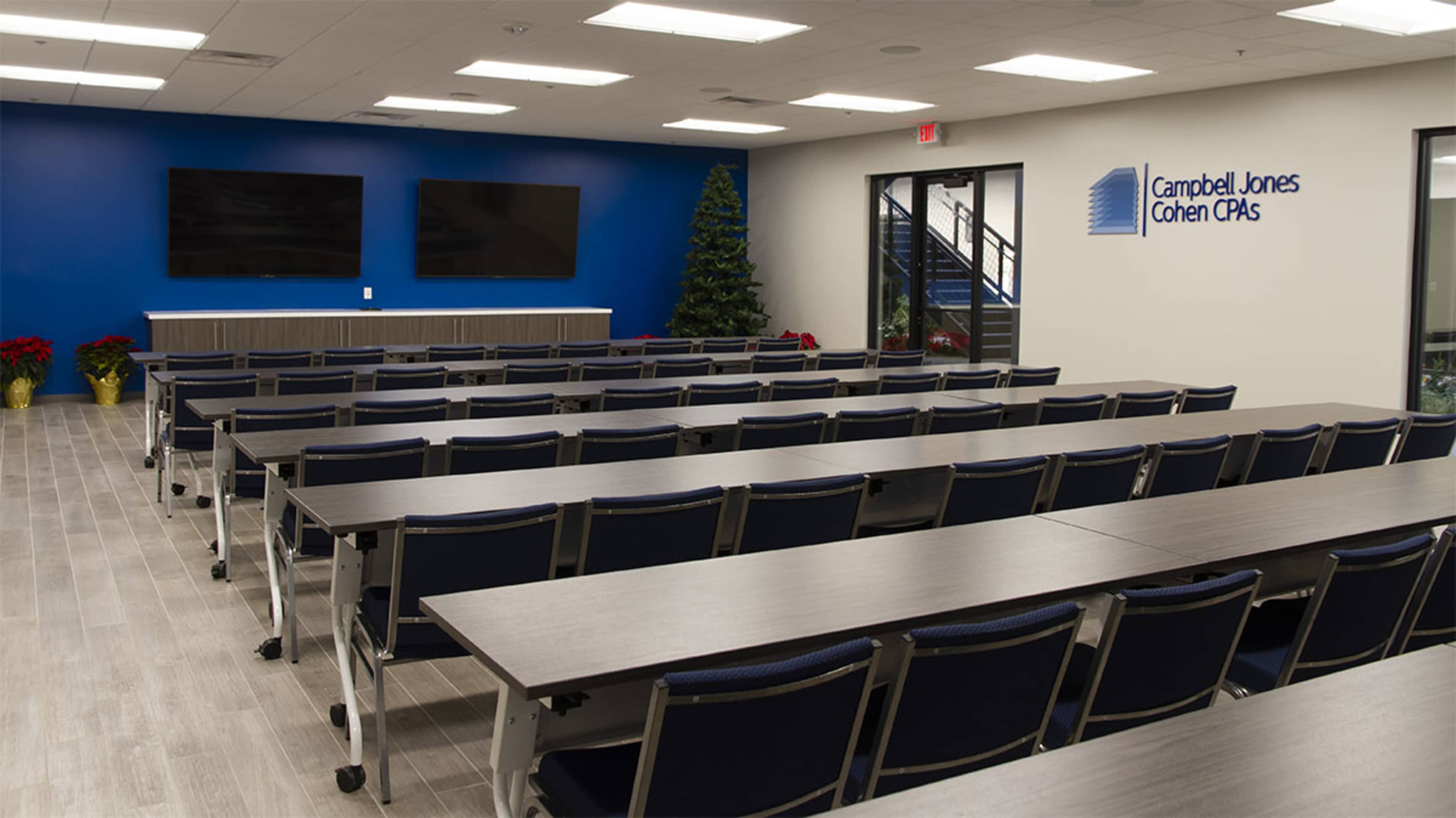 The image shows a modern, empty conference room with rows of tables and chairs facing two large screens on a blue accent wall.