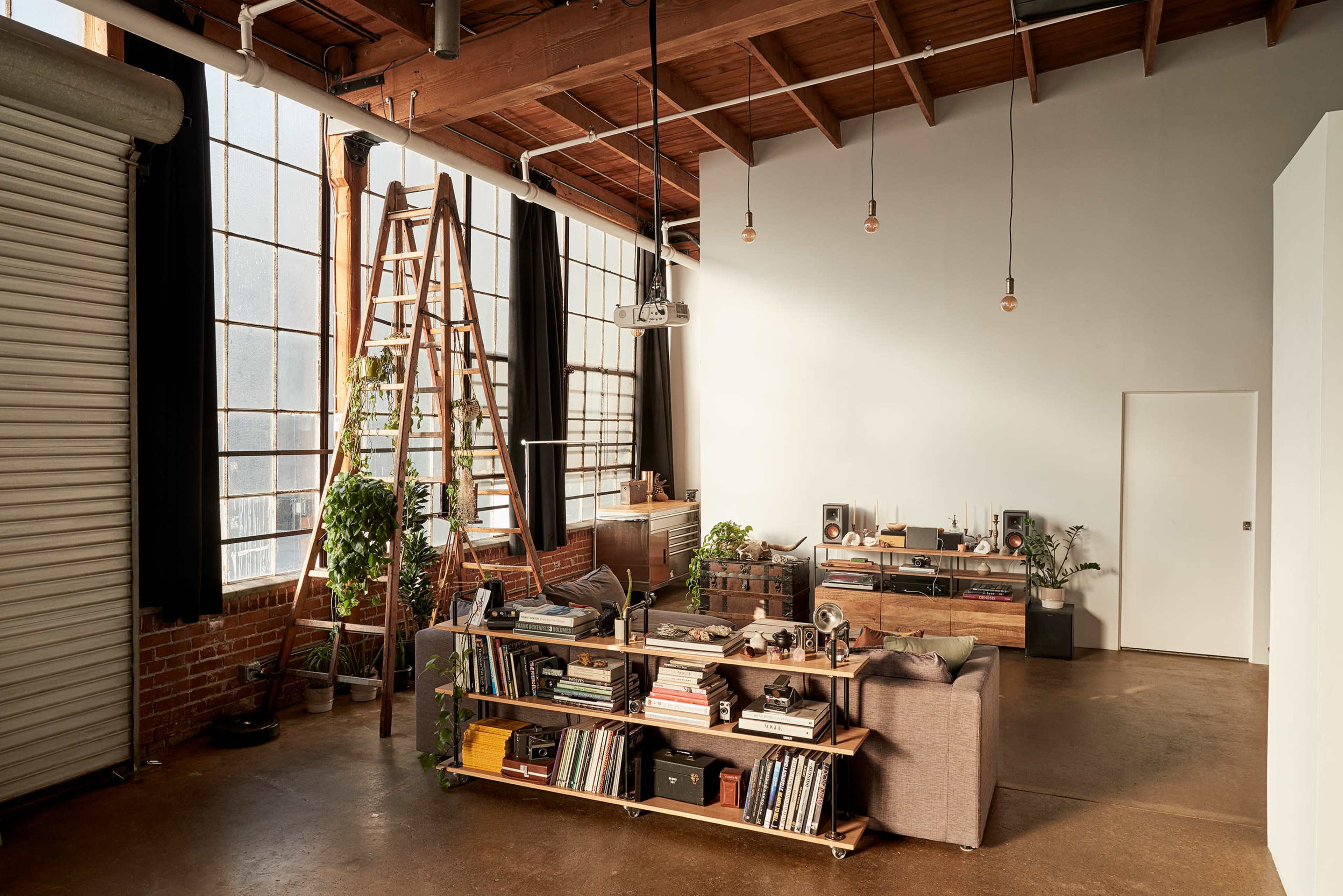A spacious room features large windows, exposed wooden beams, a ladder leaning against the wall, and various books and plants arranged on shelves and furniture.