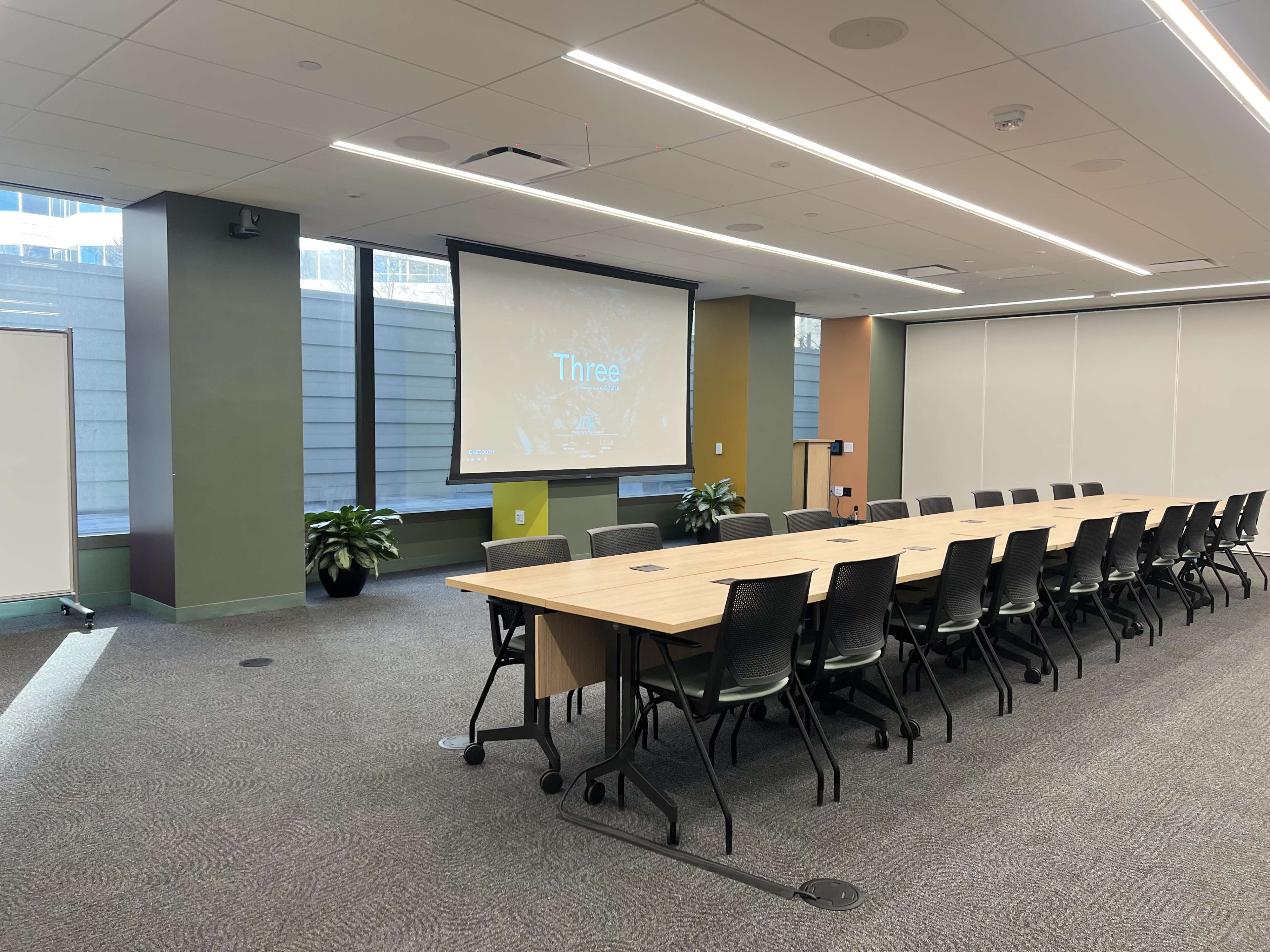A large meeting room features a long table surrounded by chairs, with a projector screen displaying the word "Three" and large windows allowing natural light in.