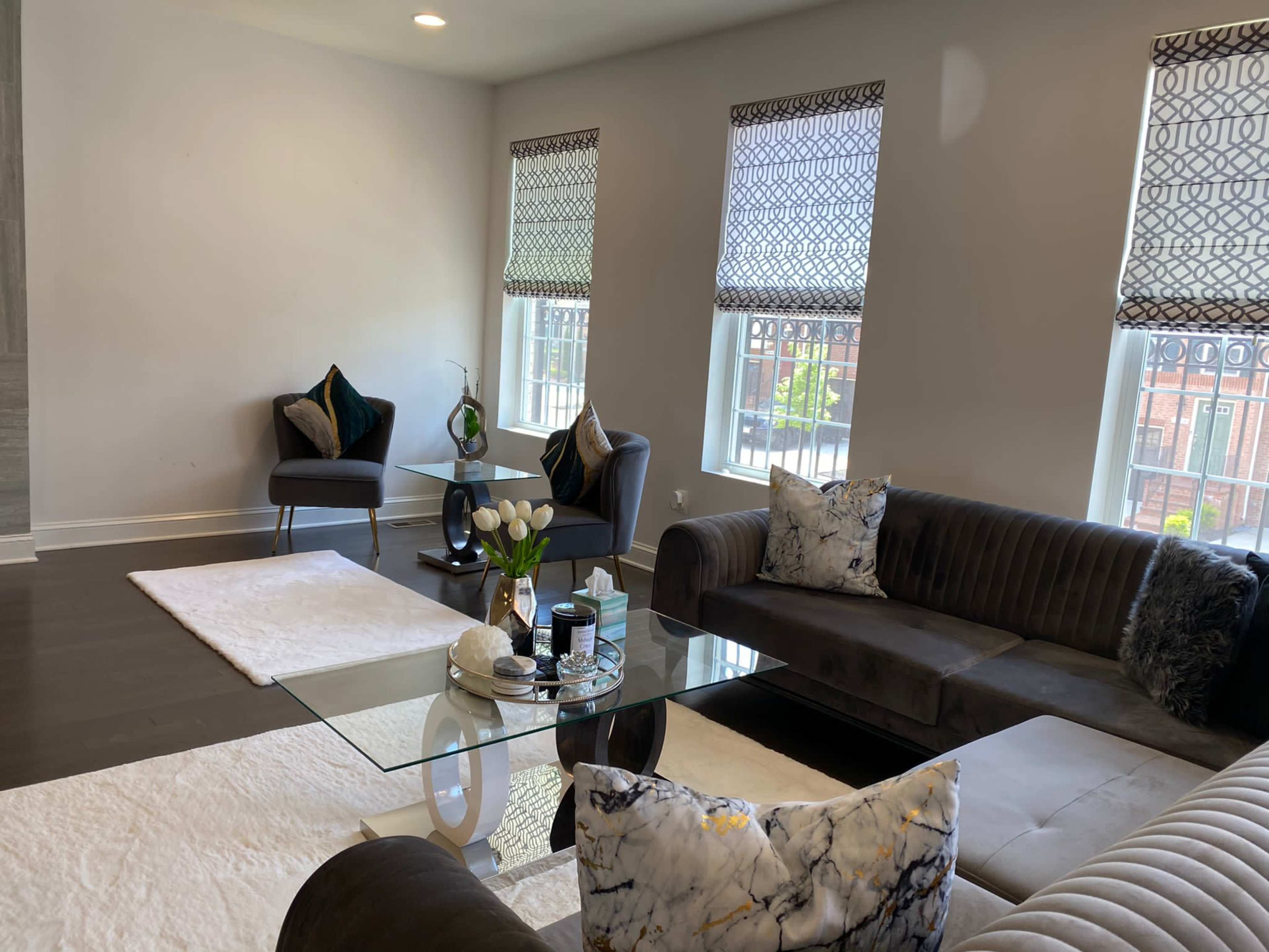 A modern living room with a glass coffee table, dark sofas, and large windows dressed with patterned shades.