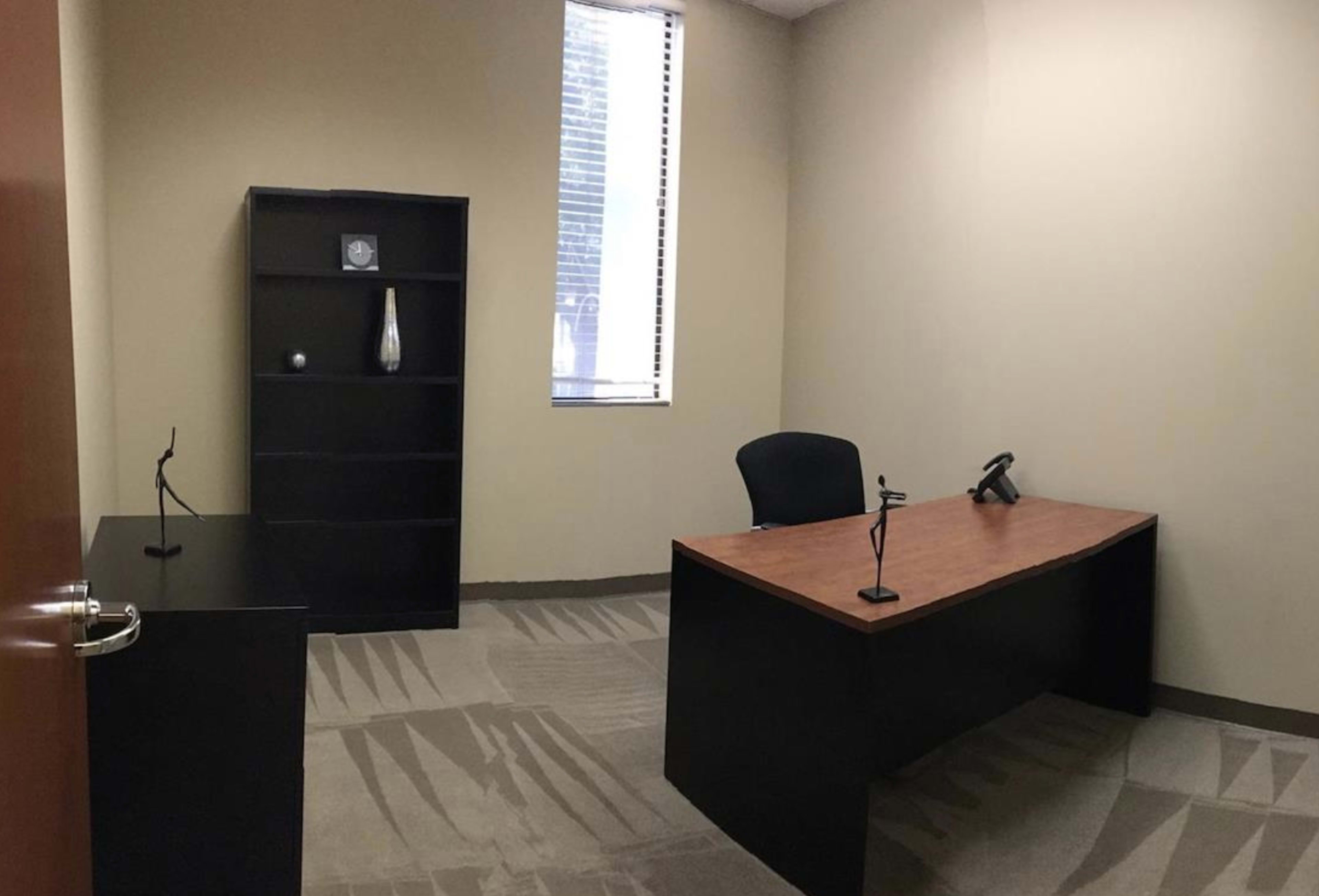 The image shows a sparsely furnished office with a black desk and a chair, a shelf against the wall, and a window allowing natural light.