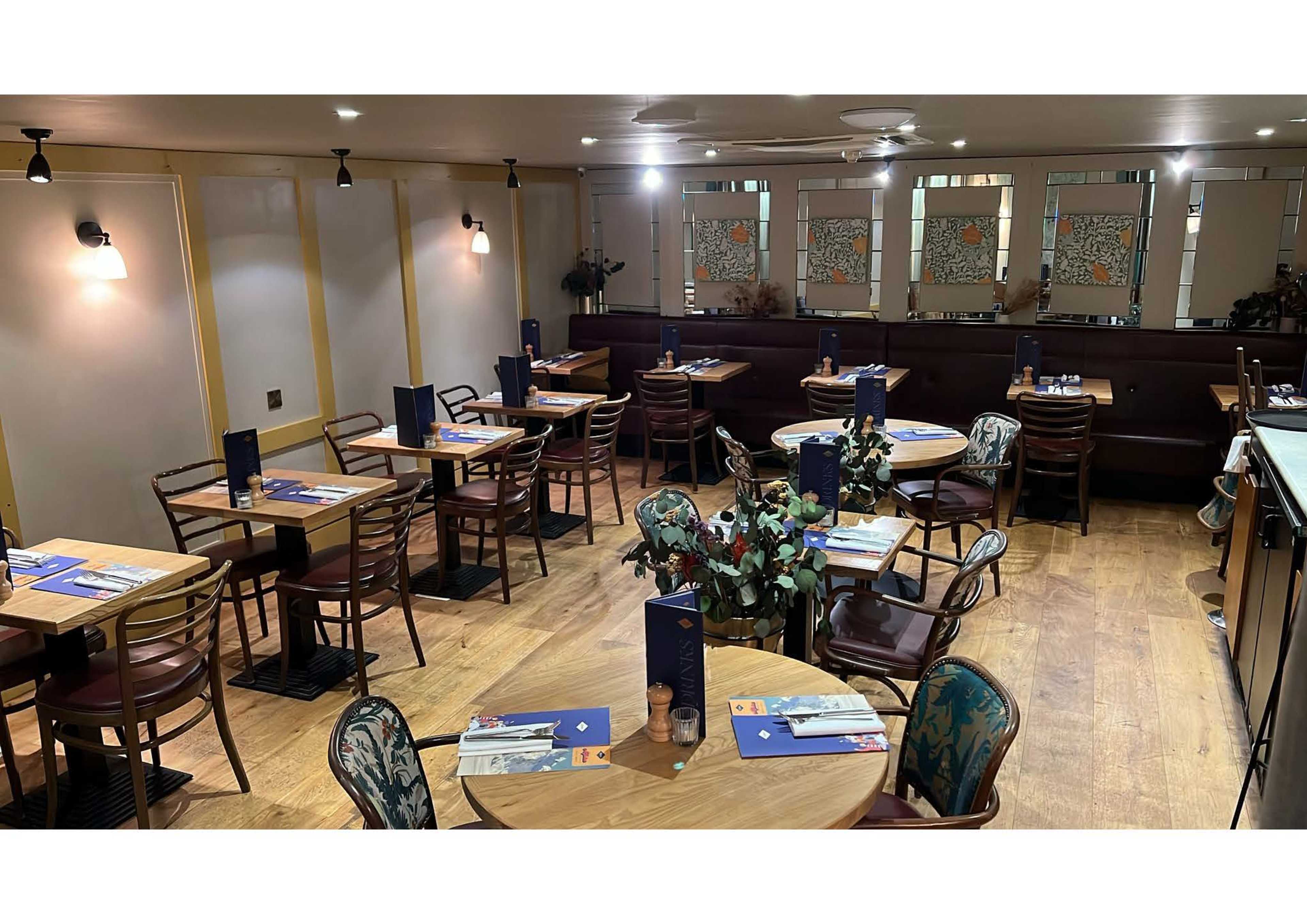 The image shows a neatly arranged restaurant interior featuring wooden tables and chairs, with decorative plants and menu stands at each table.