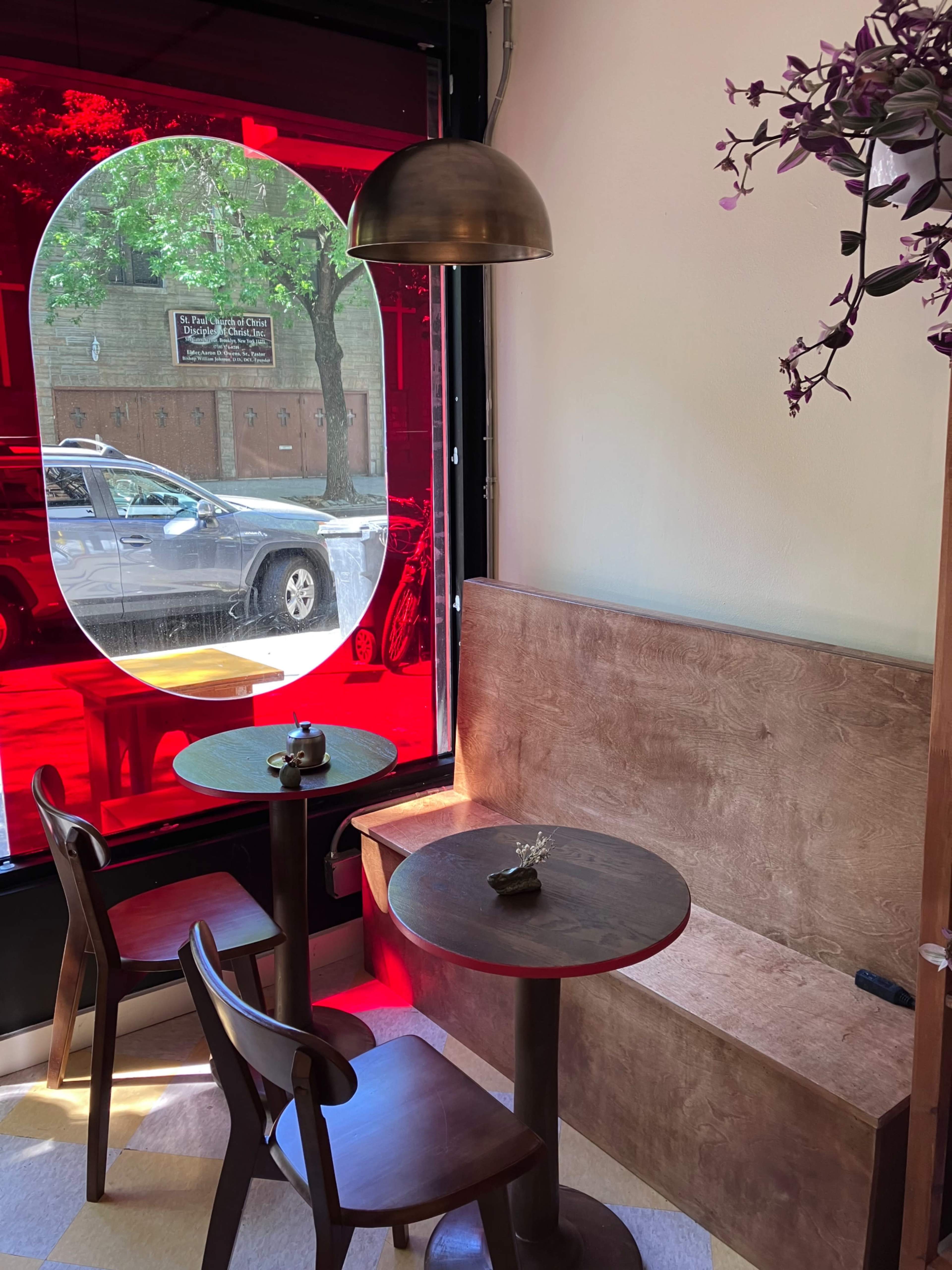 A small, modern café interior features a wooden table and chairs beside a large red-tinted window.
