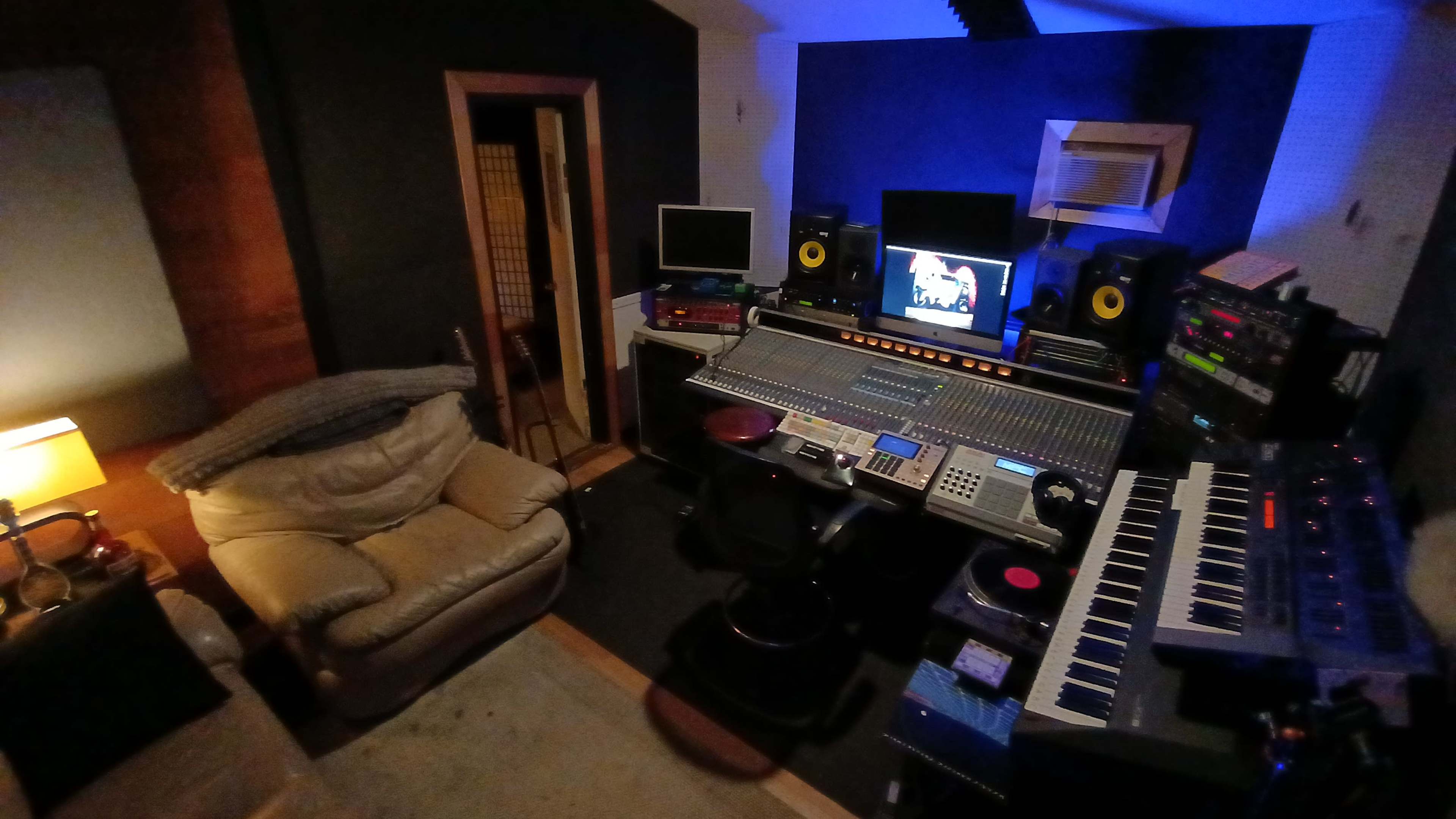 The image shows a compact music studio featuring a large mixing console, multiple computer monitors, studio speakers, and a comfortable armchair in the corner.
