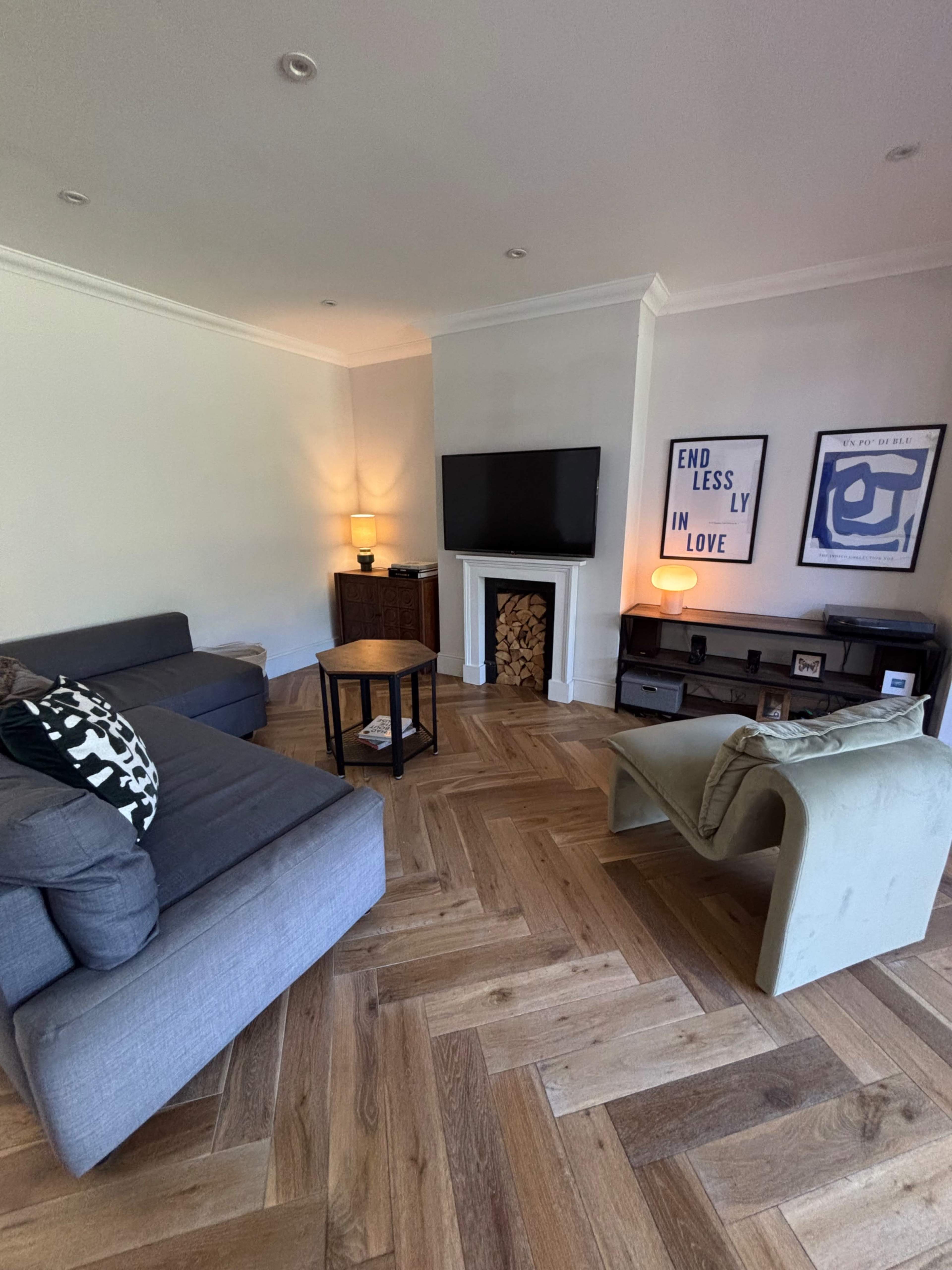 A modern living room features a gray sofa, a small round table, a television mounted on the wall, and a decorative fireplace with stacked wood.