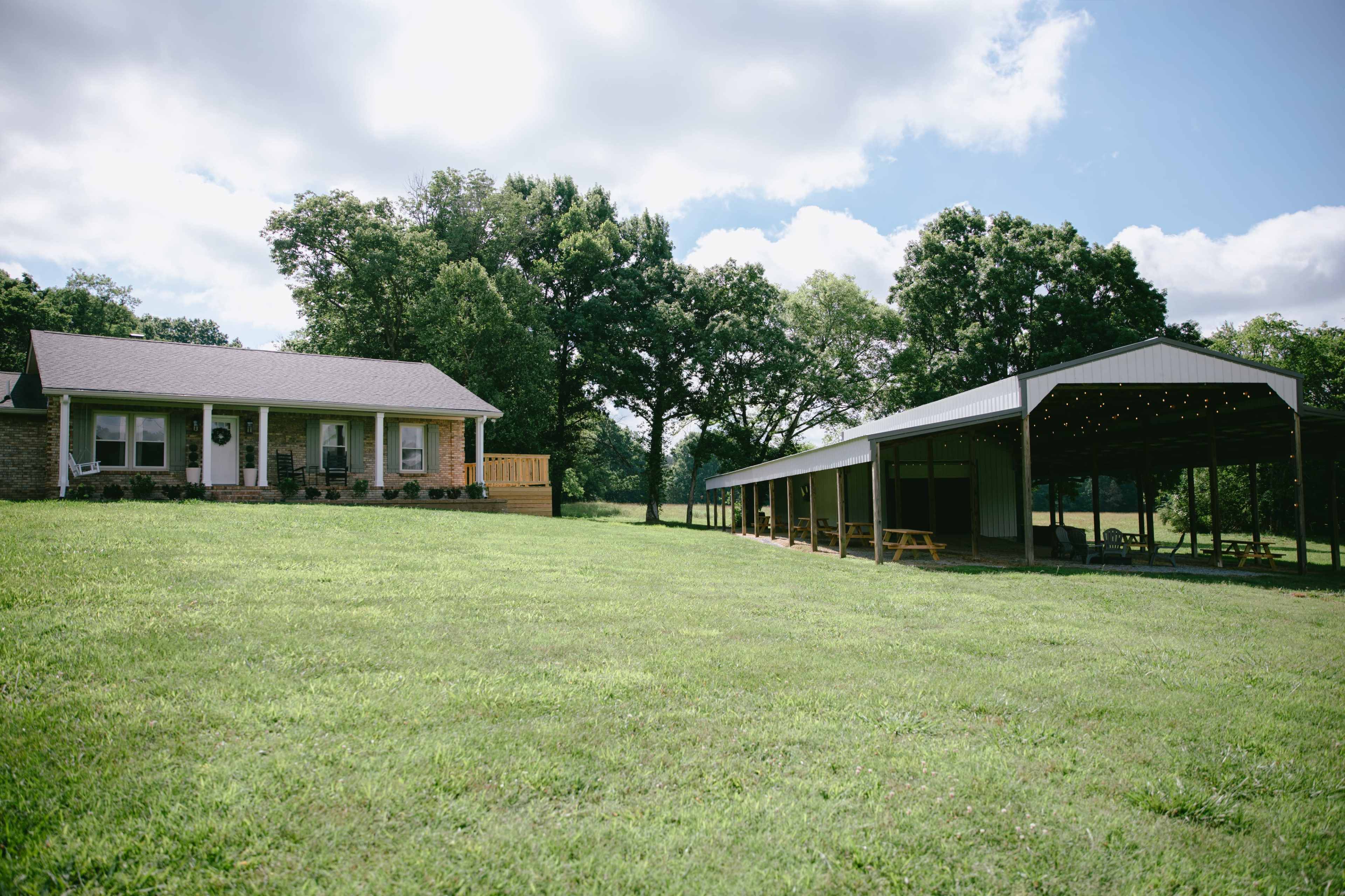 A one-story house with a front porch is situated on a grassy lawn next to a large, covered pavilion.