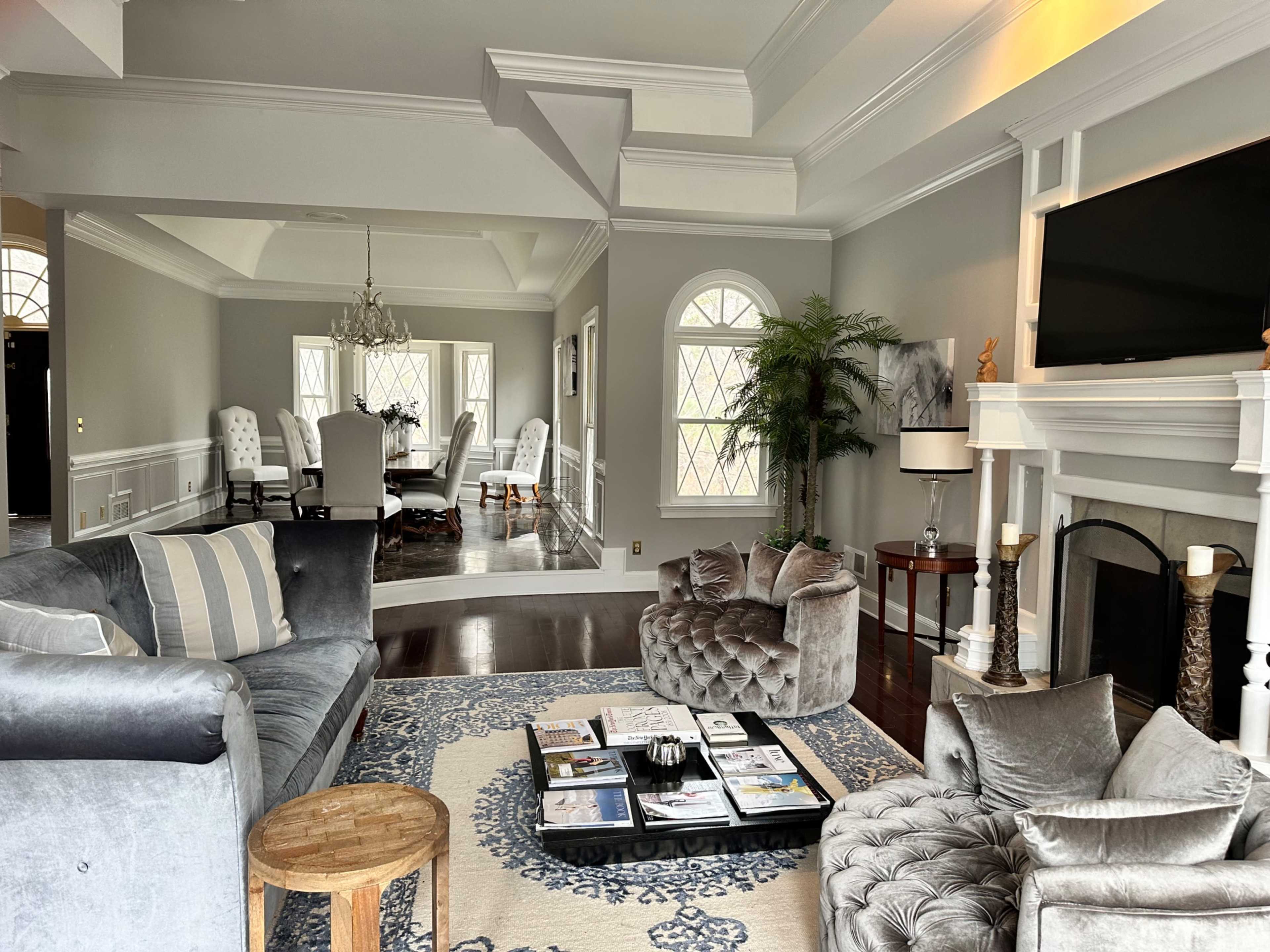 The image shows a living room with grey walls, a large sectional sofa, a coffee table with magazines, and a view into an adjacent dining area with light-colored upholstered chairs.