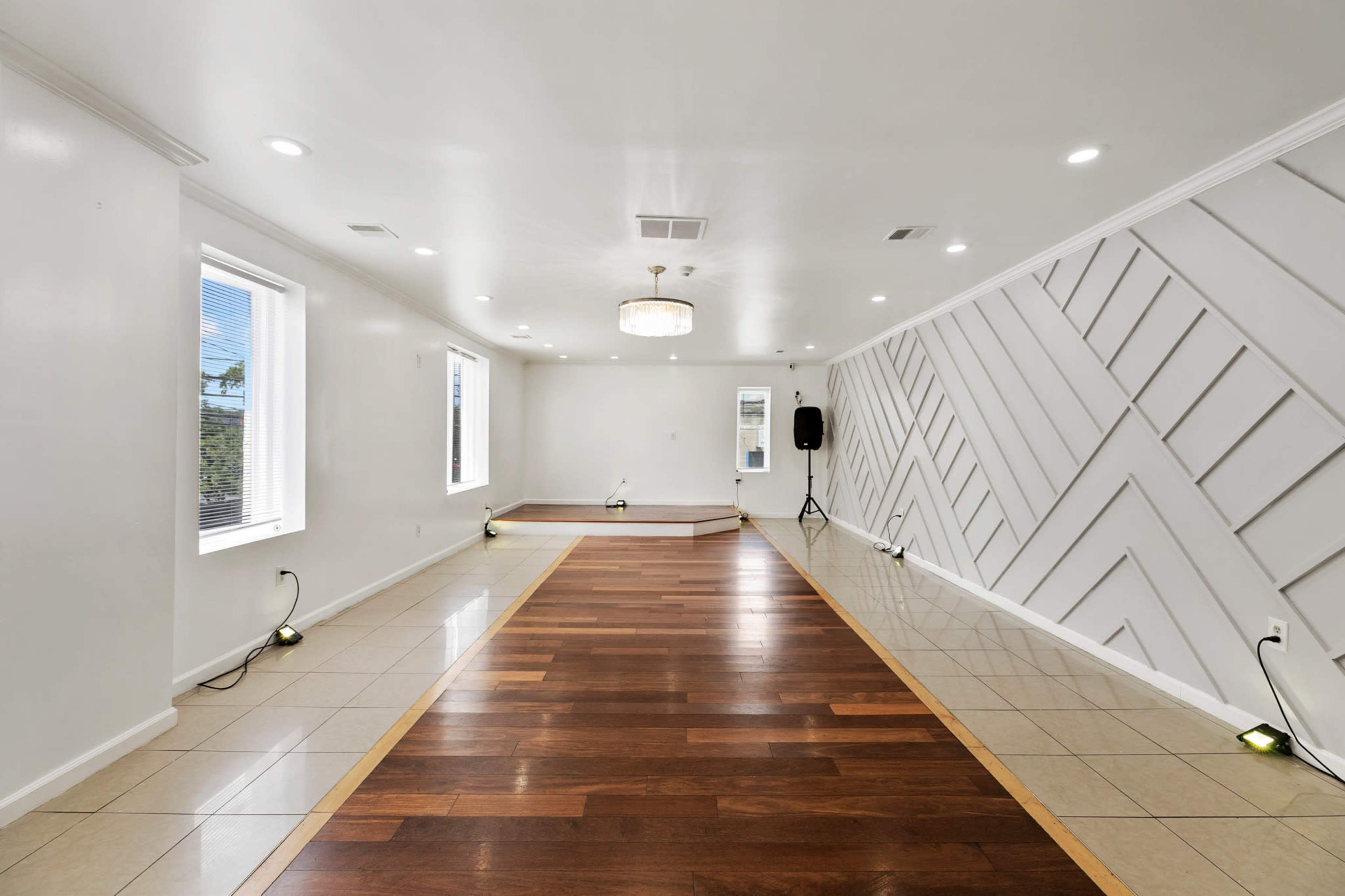 The image shows a spacious, well-lit room with a combination of tiled and hardwood flooring, decorative wall paneling, and two windows.