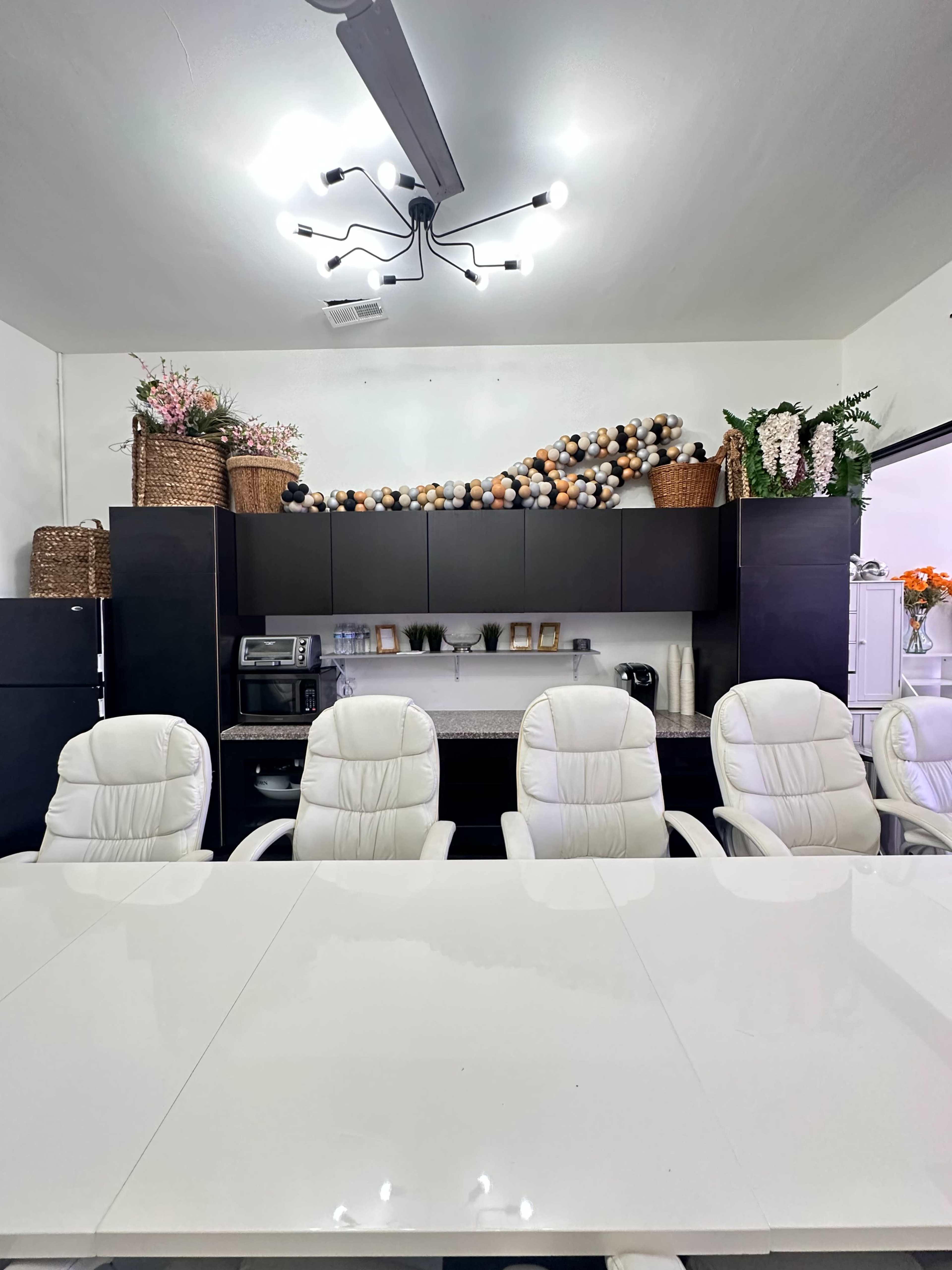 A modern conference room features a white table surrounded by several white chairs, with decorative arrangements and baskets on top of dark cabinetry in the background.