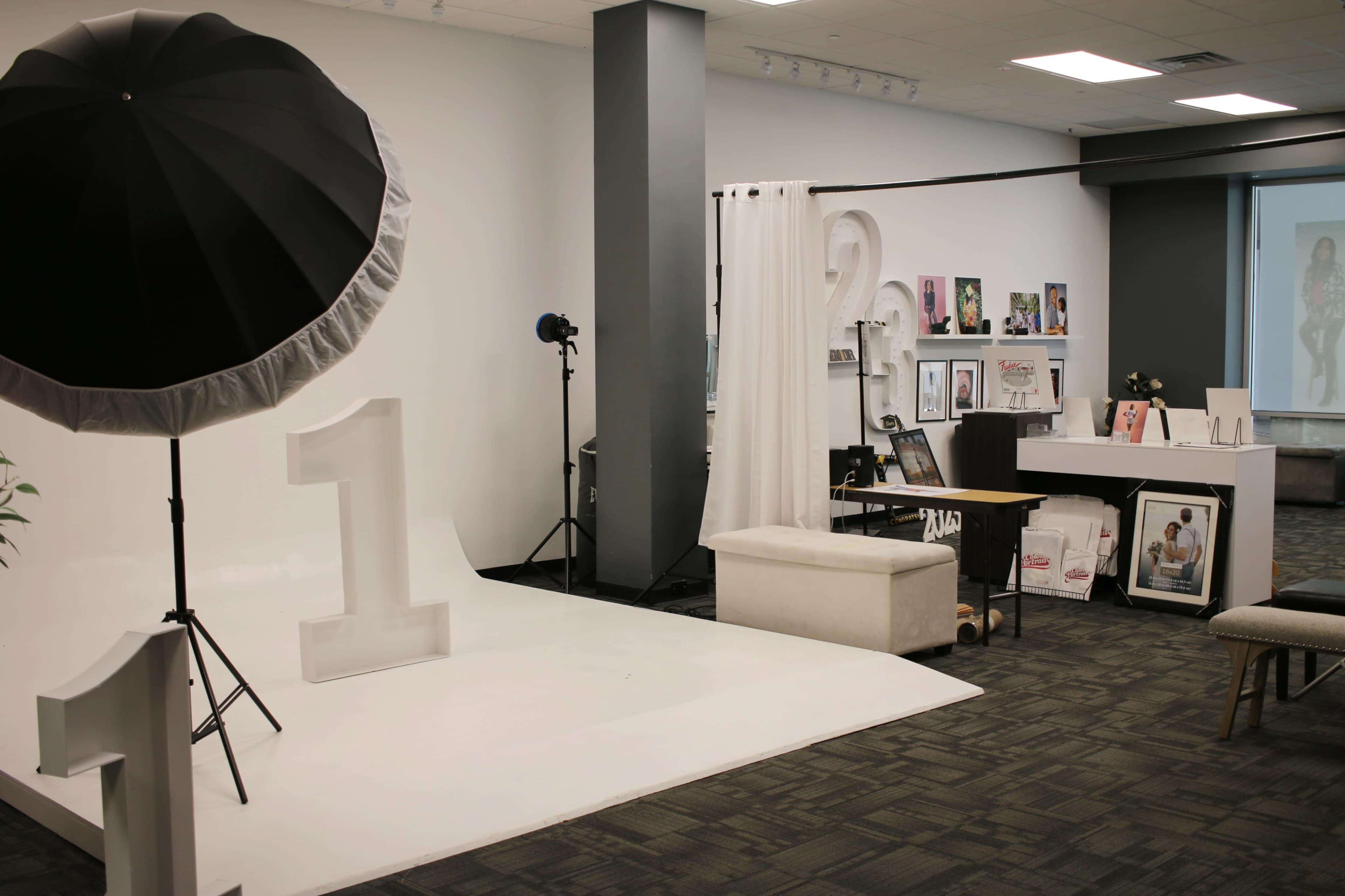 A photography studio features a white backdrop, a large umbrella light, and various displayed photo frames and magazines against a gray wall.