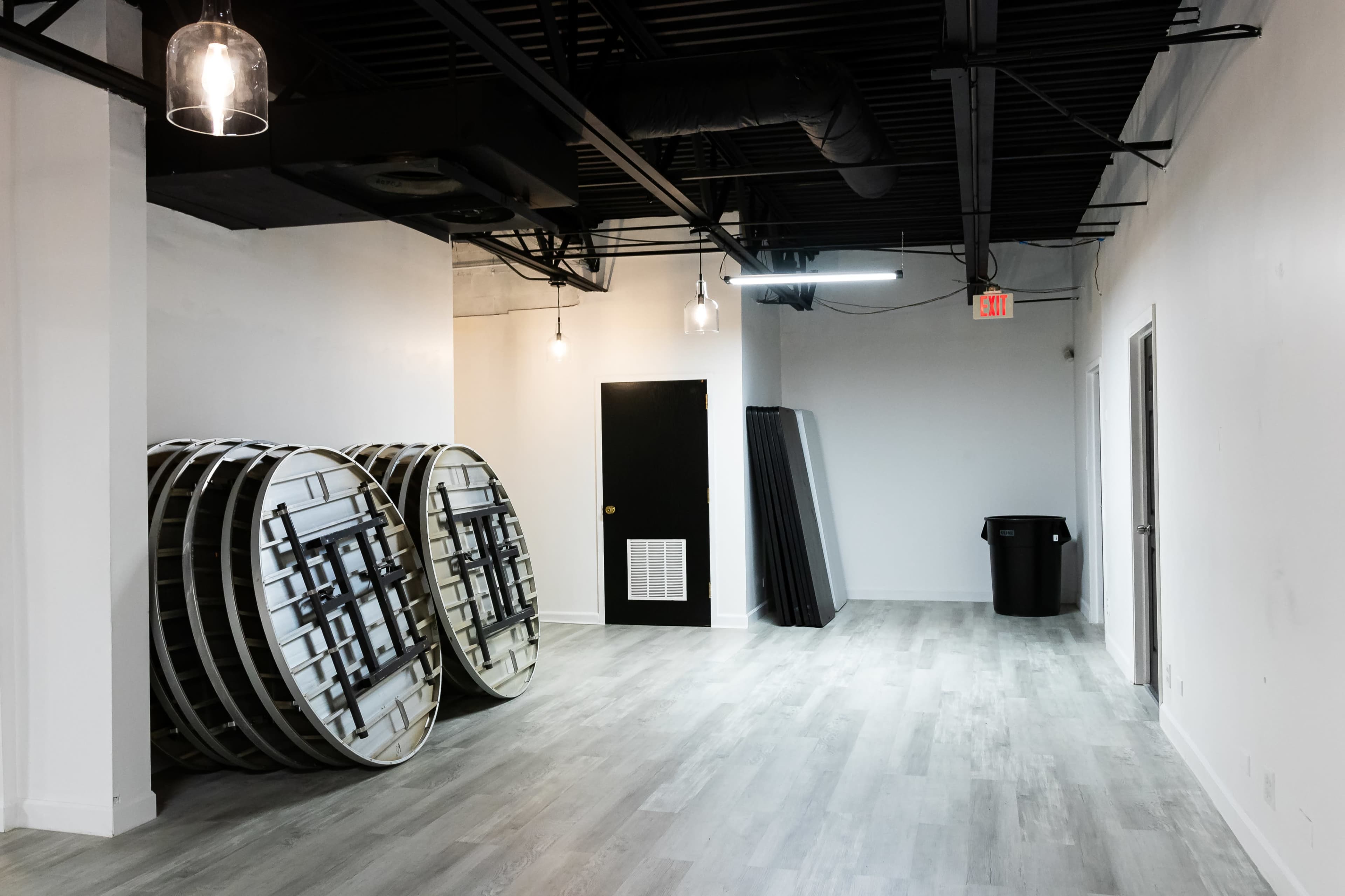 The image shows a sparsely furnished room with two large round tables leaned against a wall, a black door, a trash can, and a few other items placed against another wall.