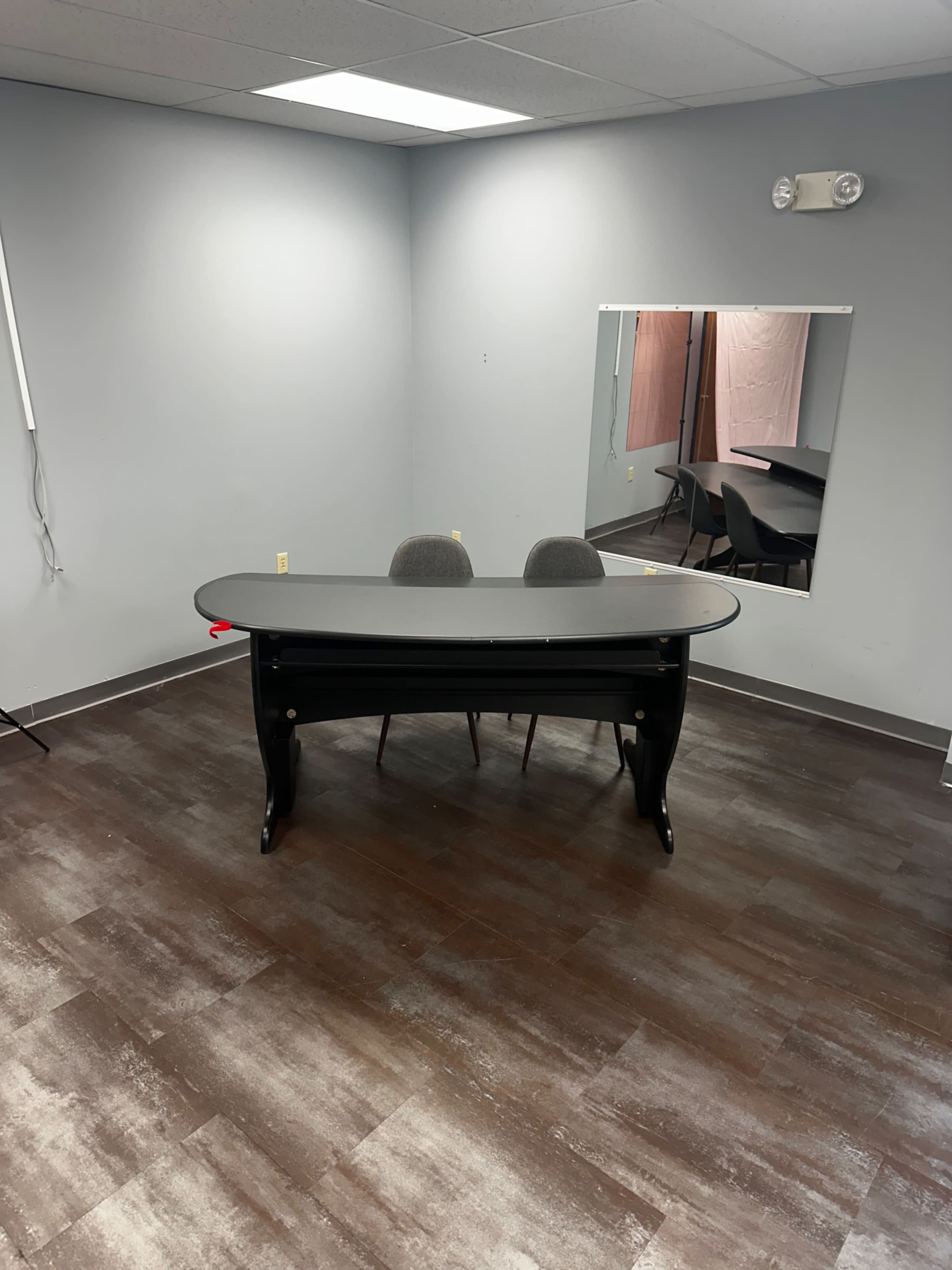 A desk with two chairs sits in an empty room that has a mirror on one wall and a light-colored background.