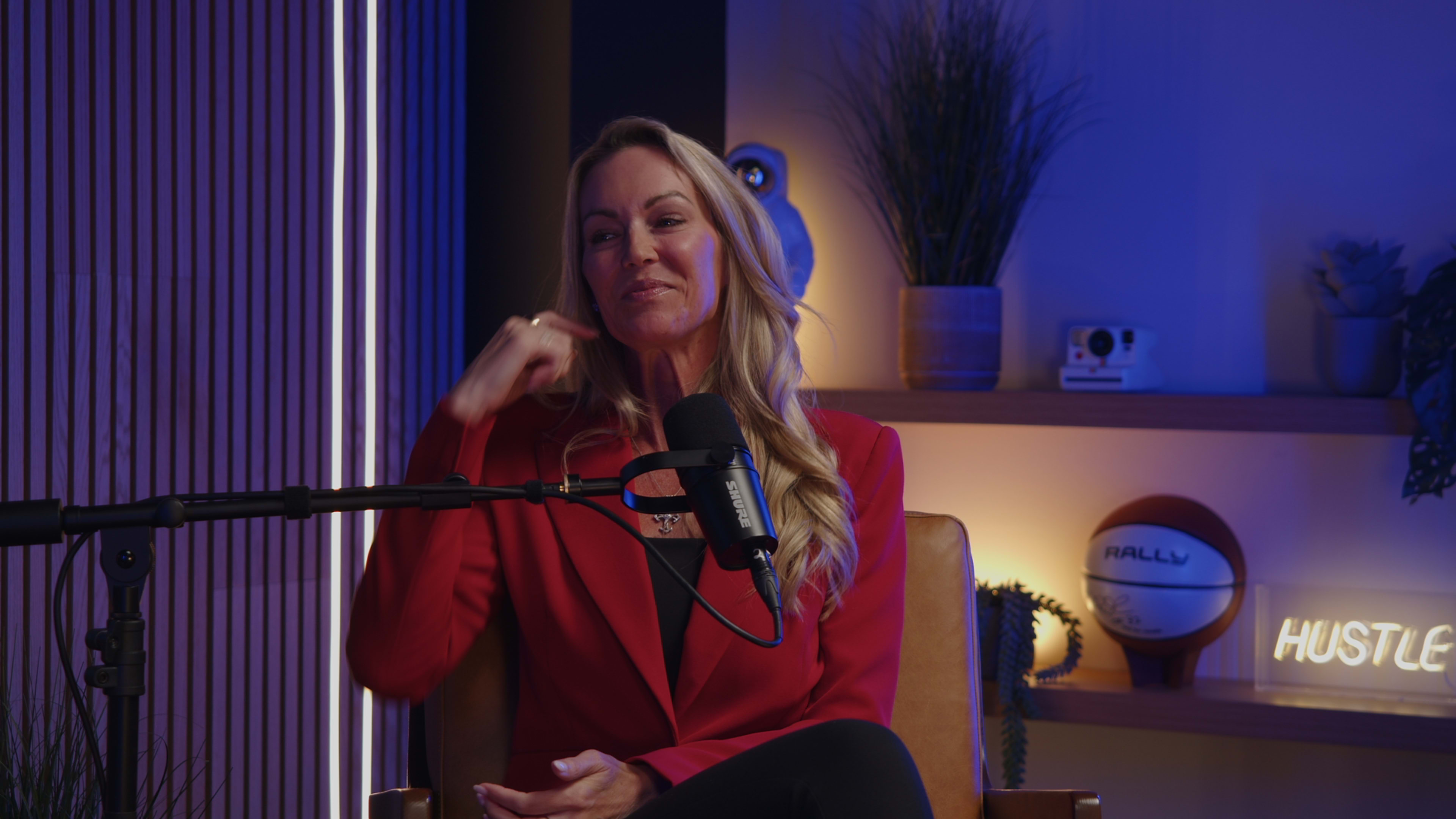 A woman wearing a red blazer sits in a chair with a microphone in front of her, surrounded by decorative plants and various objects on a shelf.