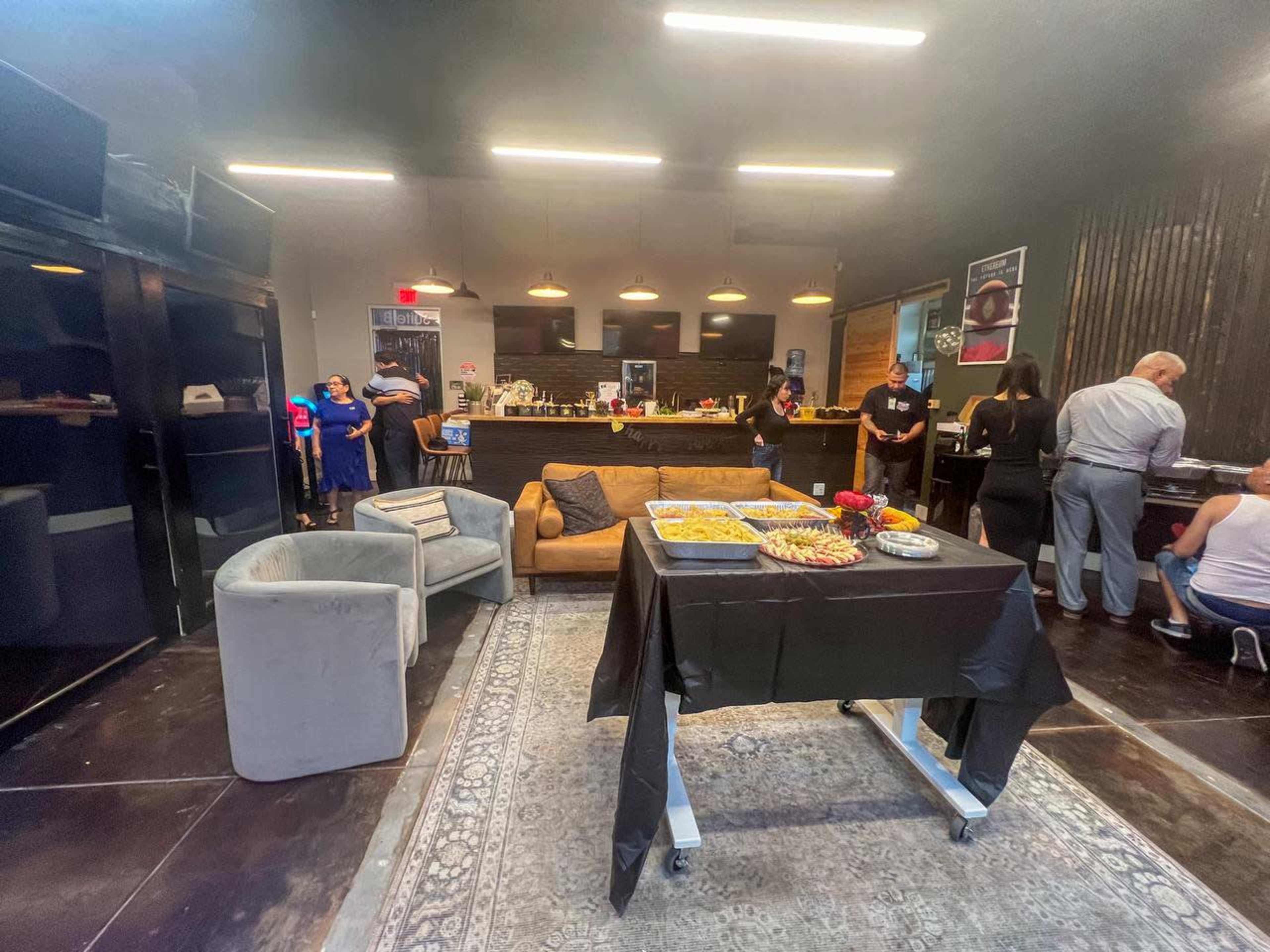 The scene depicts a spacious indoor area with a dining table covered in food, sofas, and people mingling around a bar and kitchen setup.