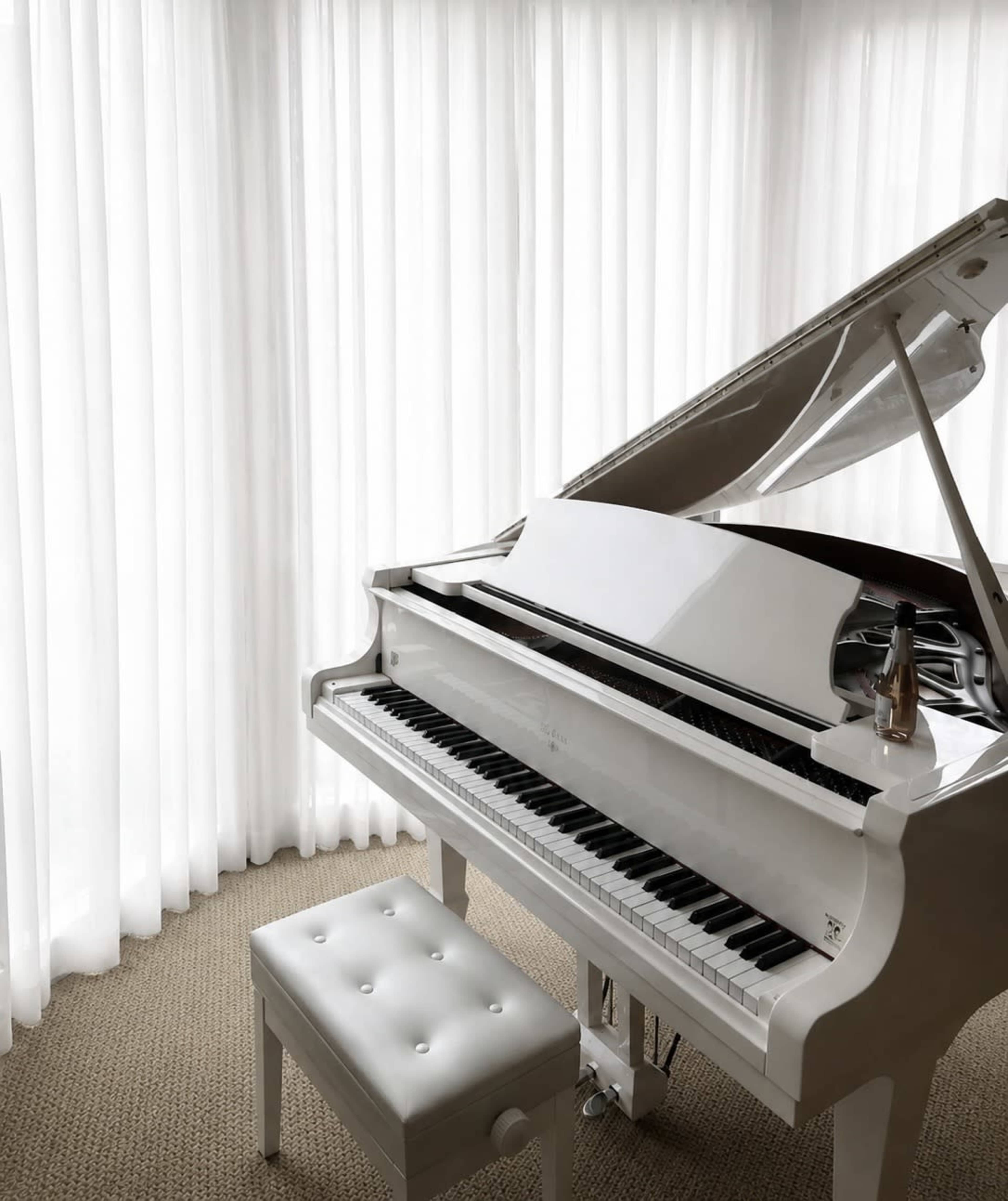 A white grand piano is positioned next to a window with sheer white curtains and a tufted white bench.