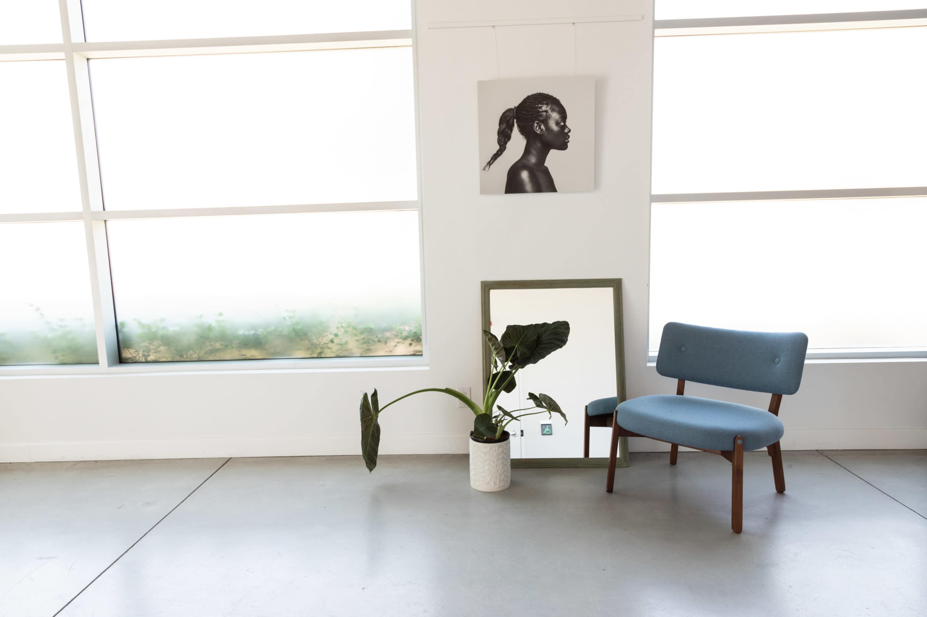 A blue chair sits next to a potted plant and a mirror, accompanied by a black-and-white portrait on the wall, with large windows in the background.