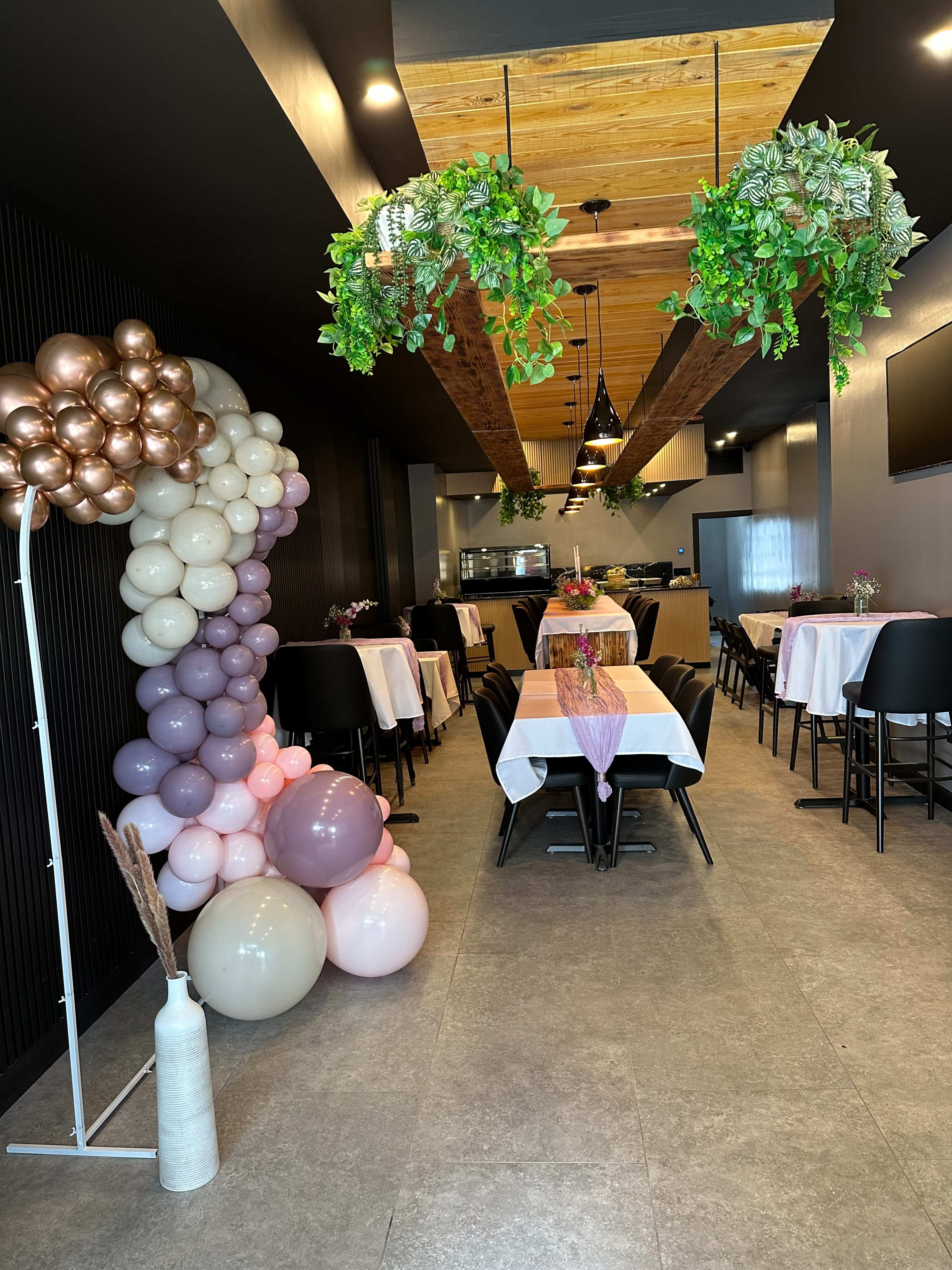 The interior of a decorated restaurant featuring tables with white tablecloths, balloon arrangements in pastel colors, and greenery hanging from the ceiling.