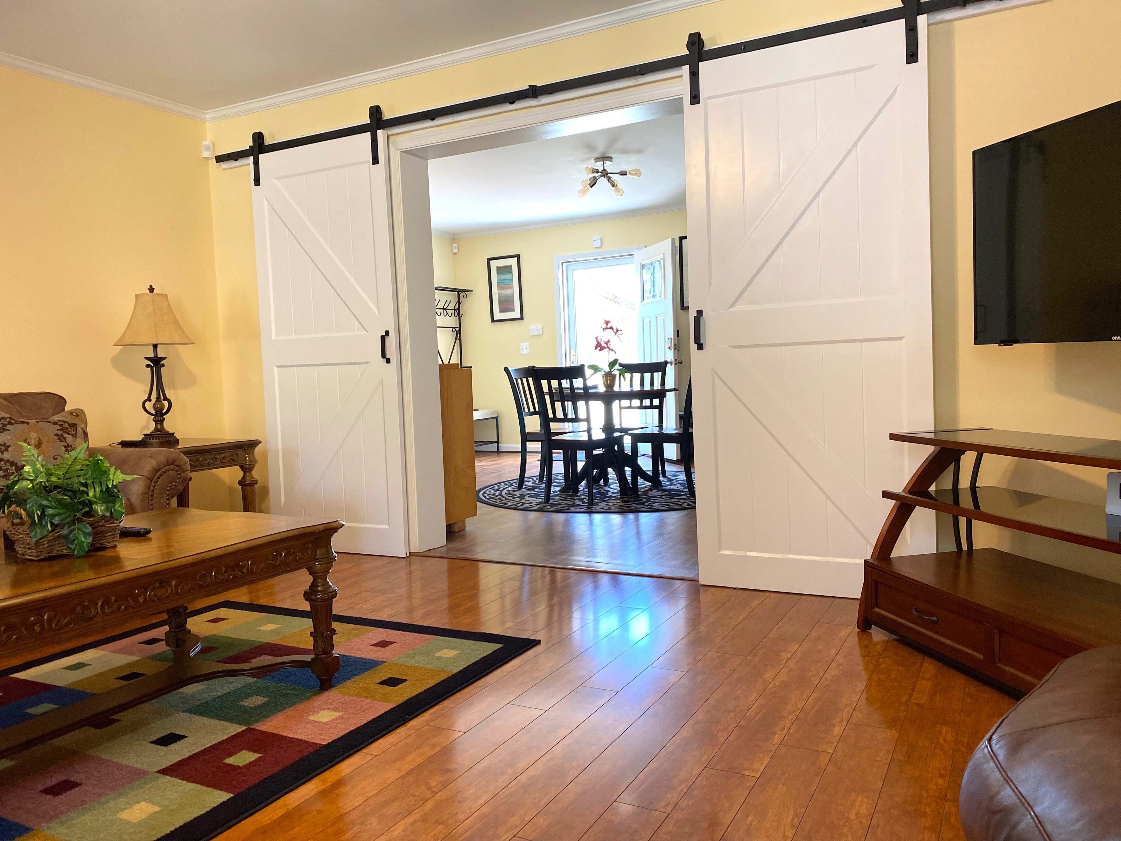 The image shows a living room with a coffee table and a couch, featuring sliding barn doors that open into a dining area with a round table and chairs.