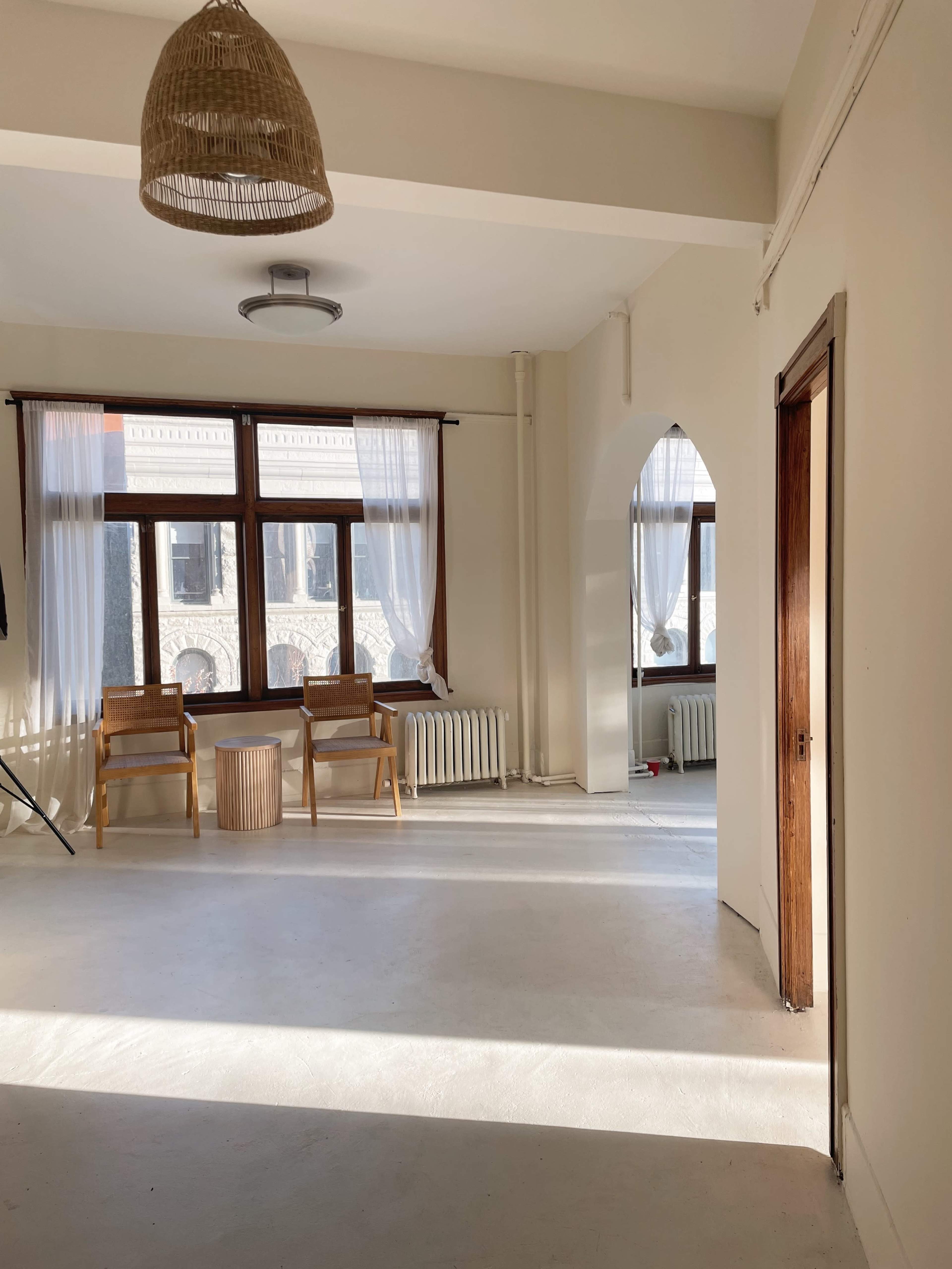 A bright, minimalist room with two wooden chairs facing a small round table, large windows with sheer curtains, and a light fixture overhead.