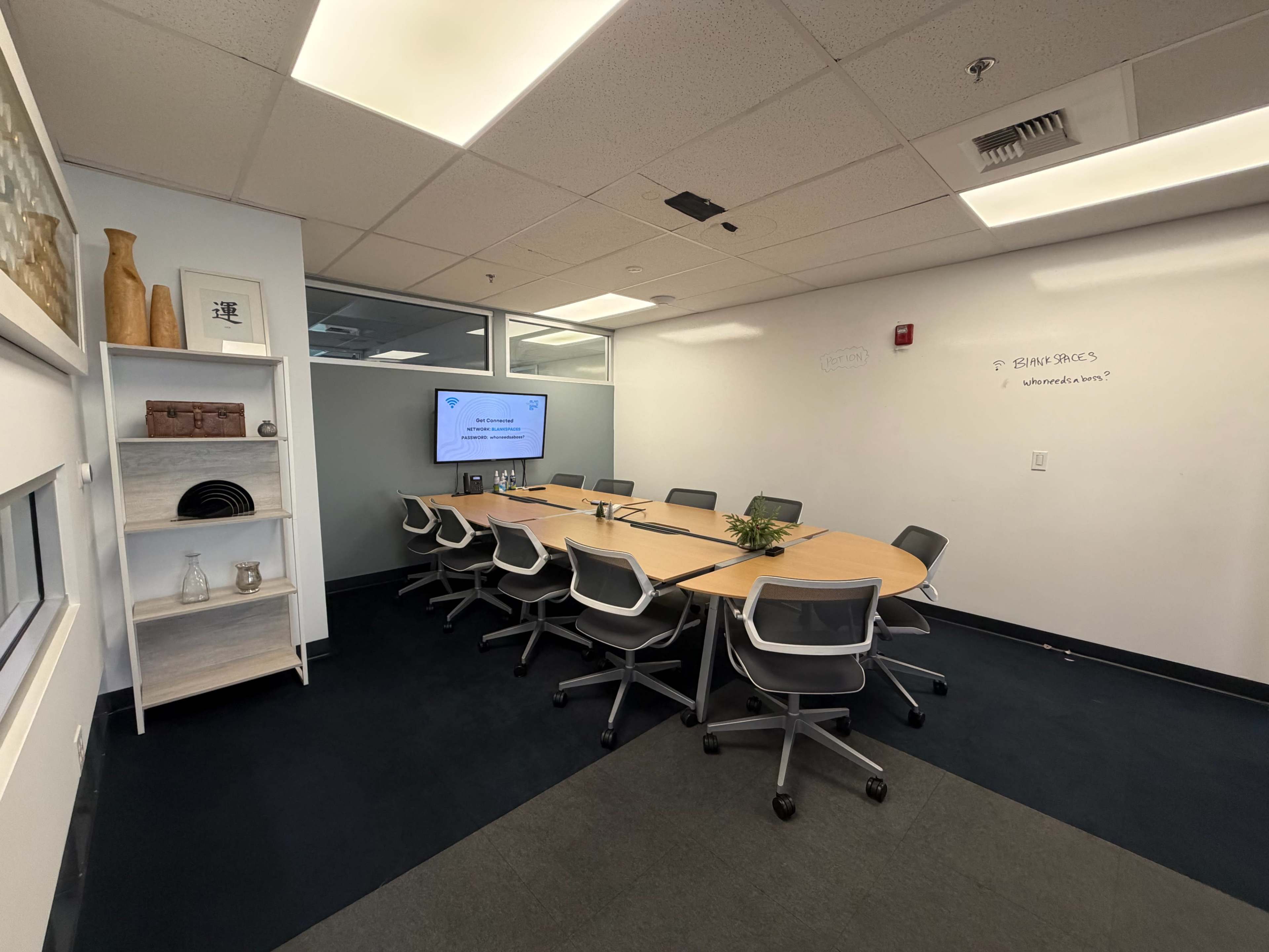 The image shows a modern conference room with a long wooden table, ergonomic chairs, a wall-mounted monitor, and a whiteboard.