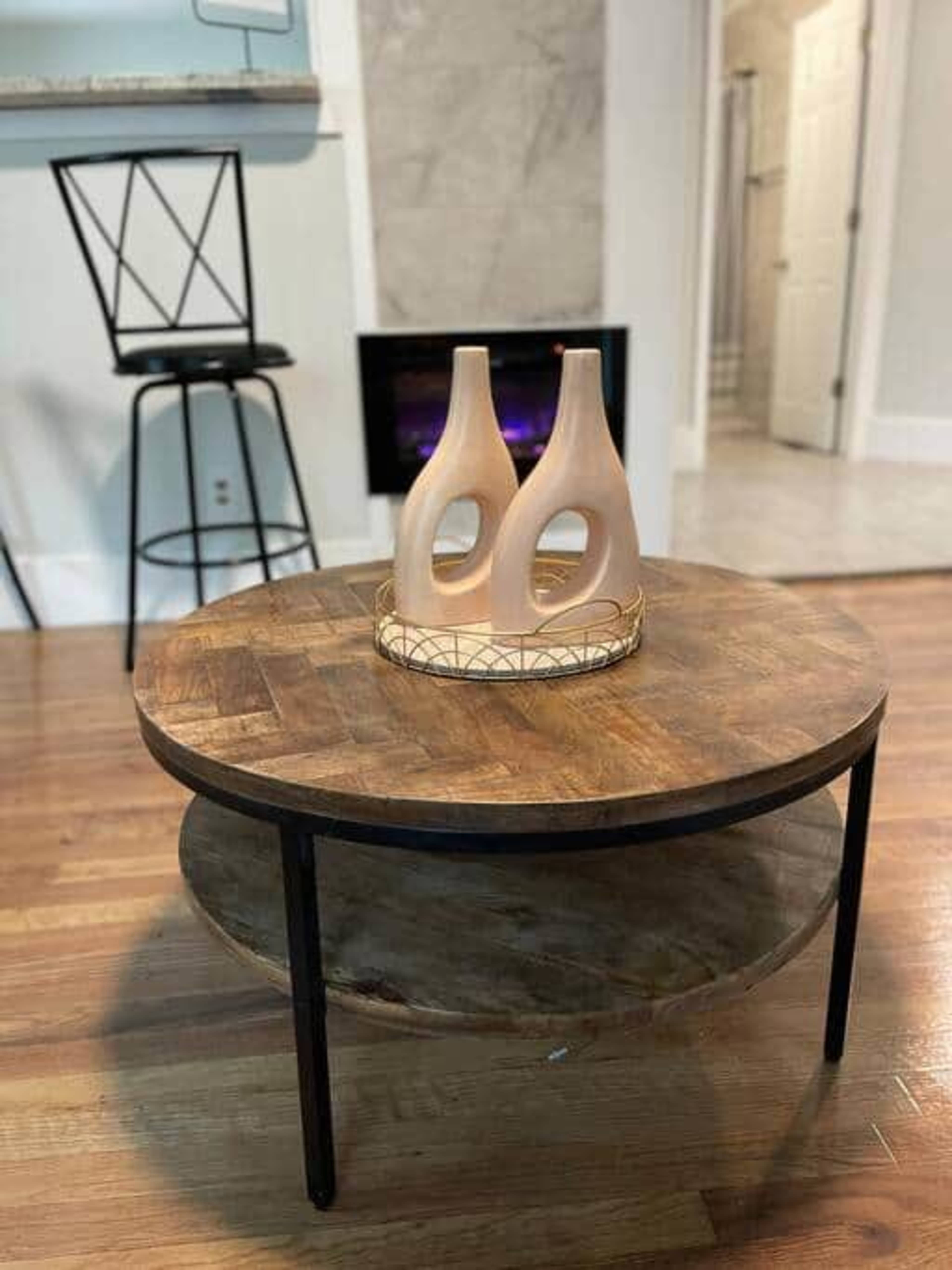 A round wooden coffee table with a lower shelf features two ceramic vases on a decorative tray, positioned in a room with a bar stool and a visible doorway to a bathroom.