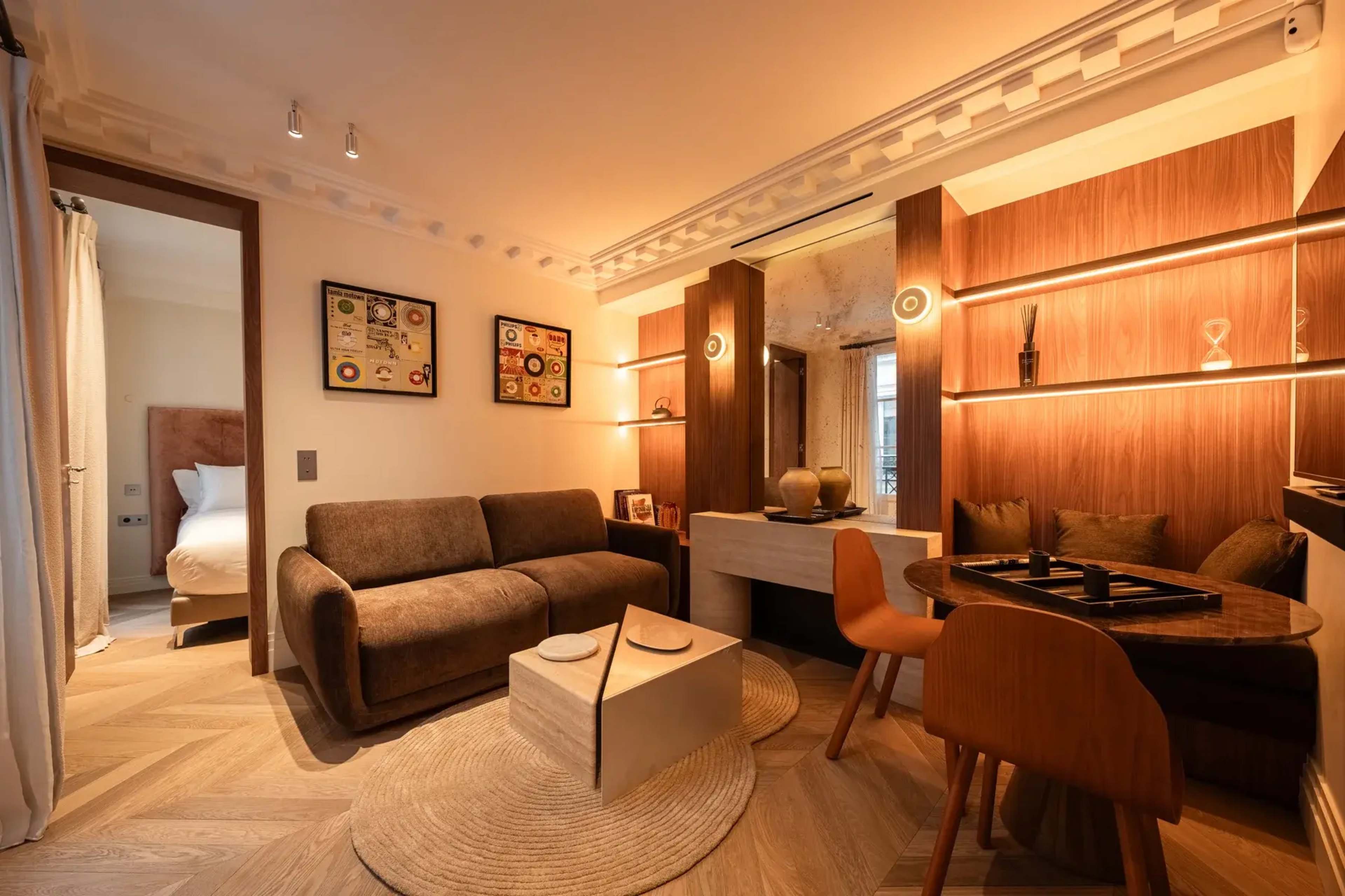 The image shows a modern living space featuring a brown sofa, a small dining table with wooden chairs, and warm lighting, all accented by decorative elements on the walls.