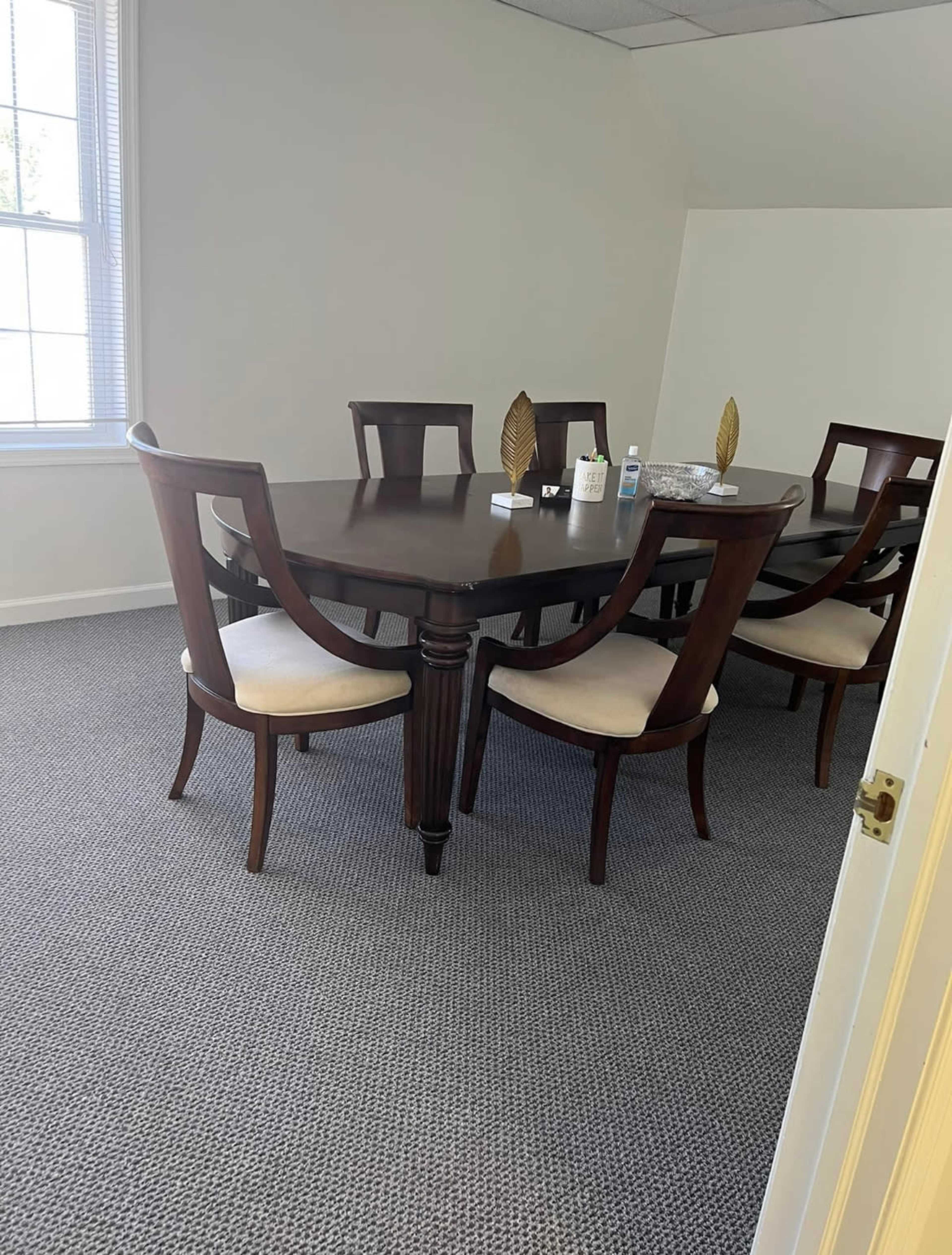A brown wooden dining table surrounded by six matching chairs is positioned in a well-lit room with beige carpet and white walls.