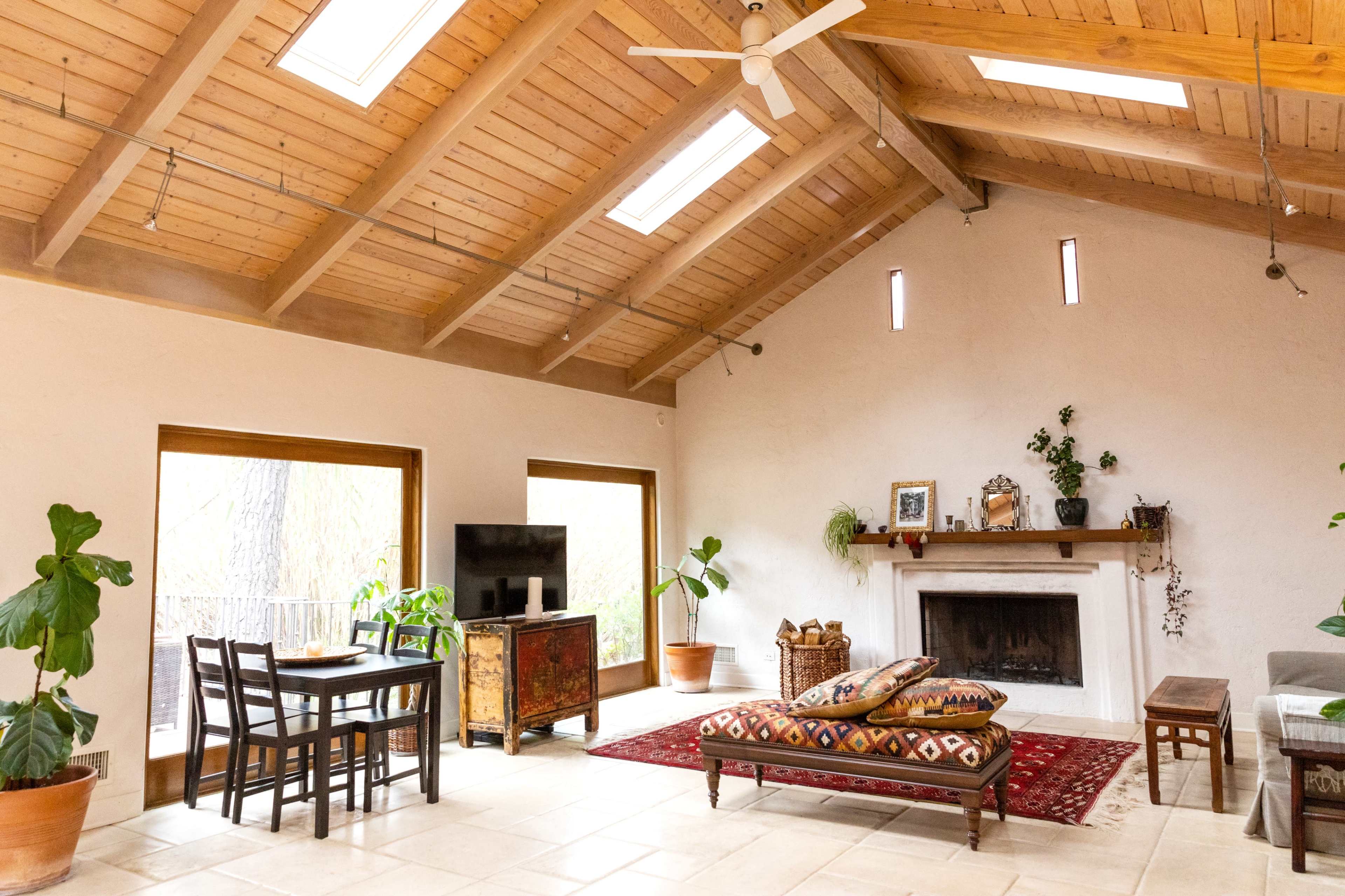 A spacious living area with a high wooden ceiling, a dining table, a television, a sofa, and a fireplace, accented by potted plants and decorative items.