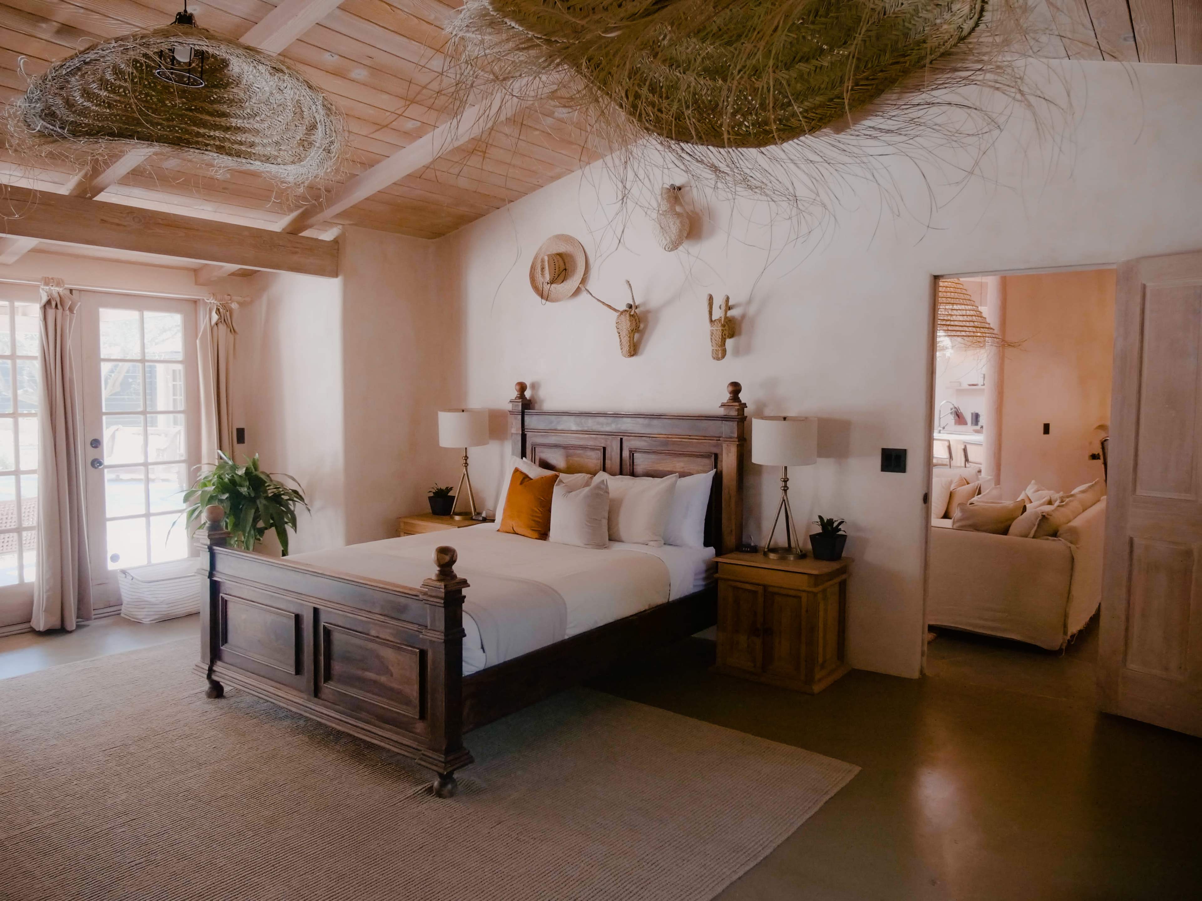 A well-furnished bedroom features a wooden bed, two lamps on nightstands, and natural decor elements, with an adjacent cozy sitting area visible through an open doorway.