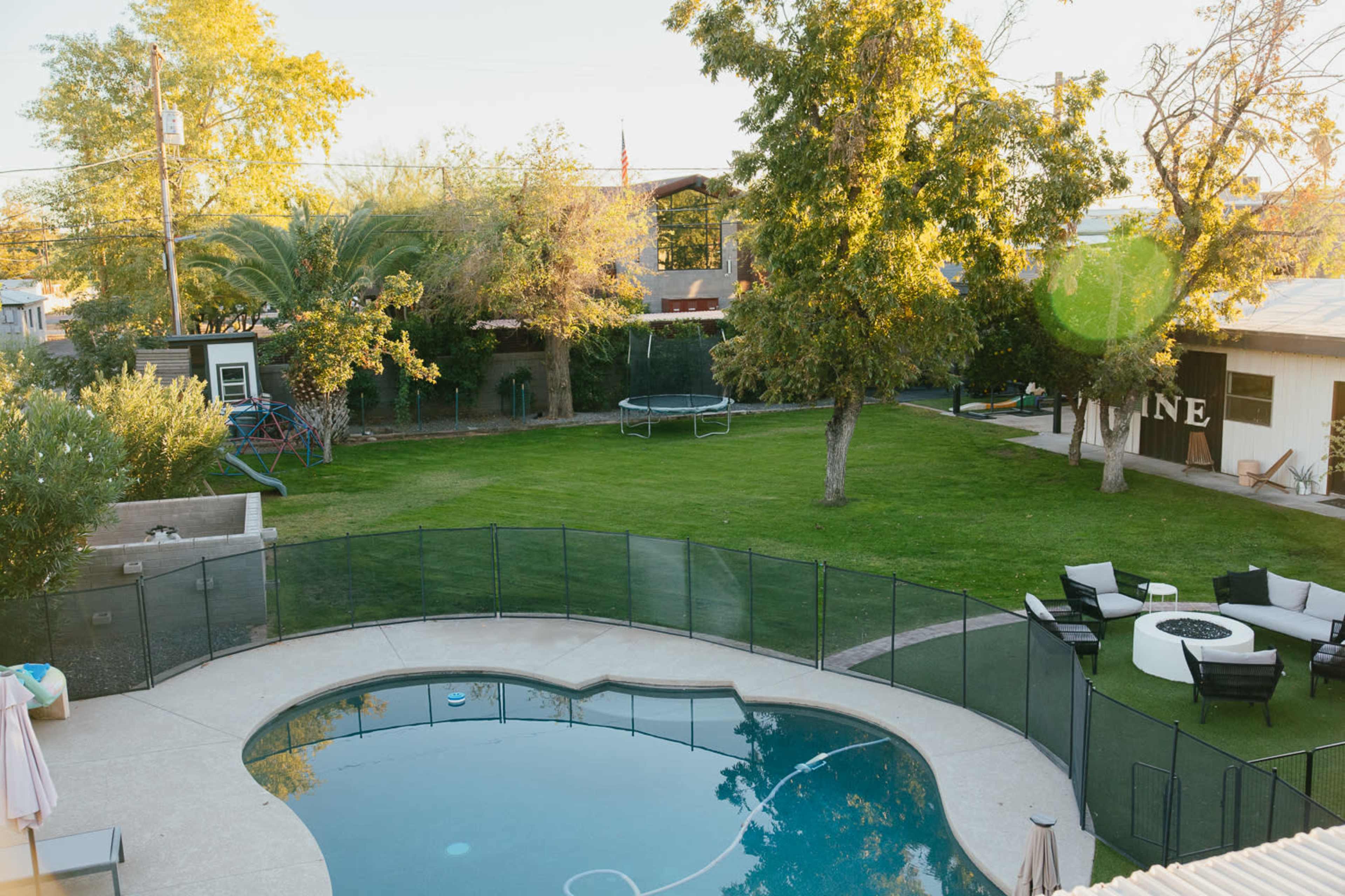 A backyard with a swimming pool, a fenced yard, a trampoline, and seating areas surrounded by trees.