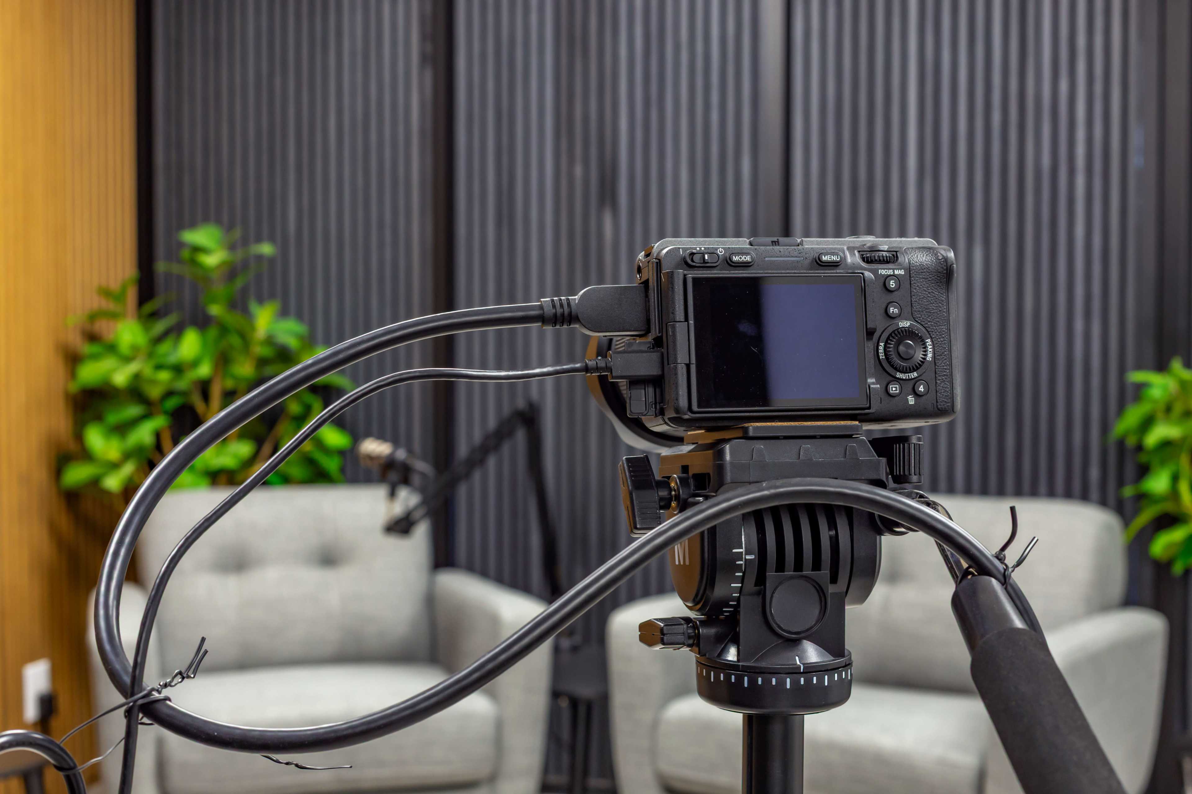 A camera is mounted on a tripod with various cables connected to it, overlooking two empty chairs against a backdrop of vertical wood paneling and greenery.