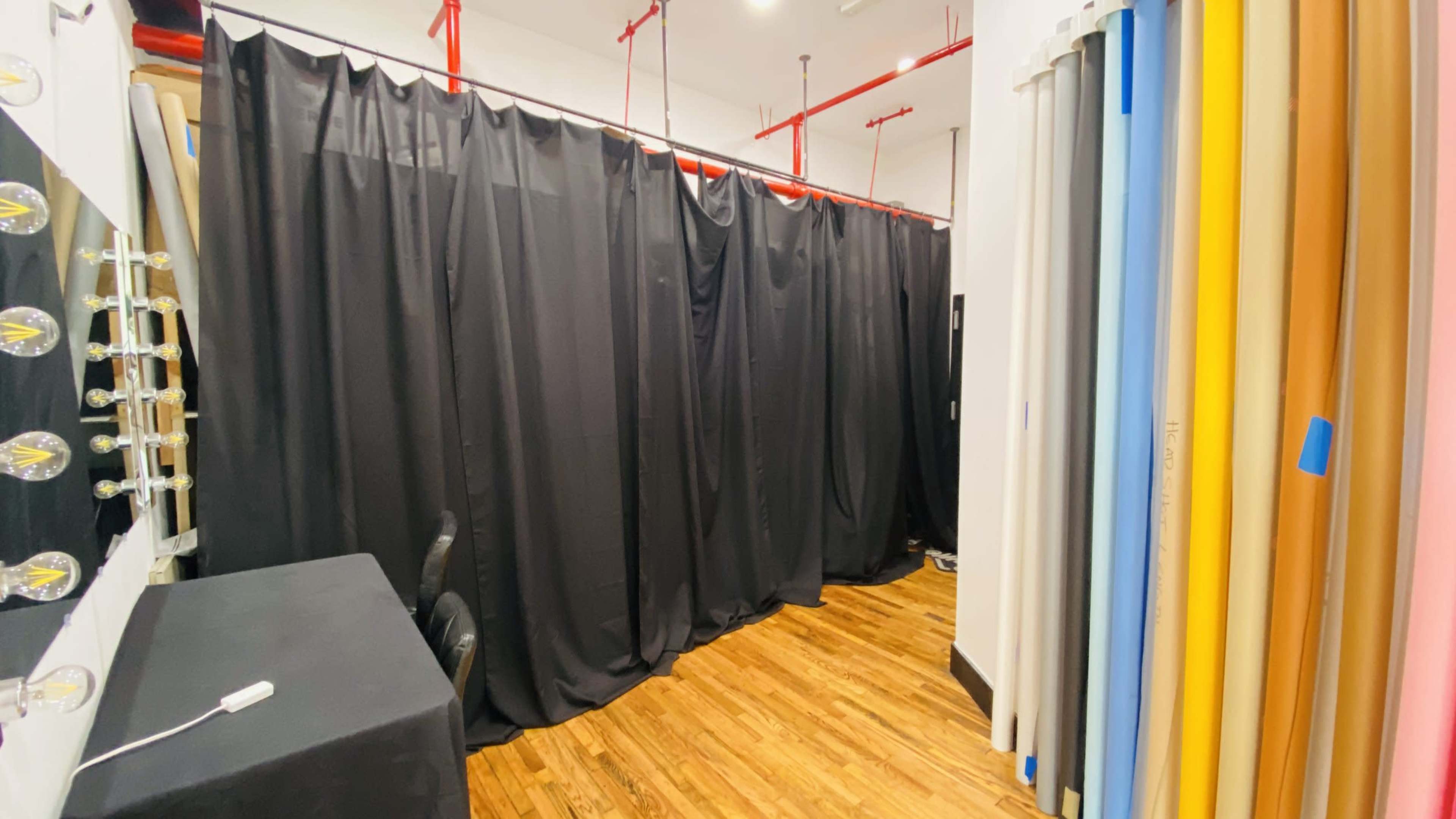 The image shows a studio space with black curtains, a table and chairs, and various backdrop rolls in pastel colors along one wall.