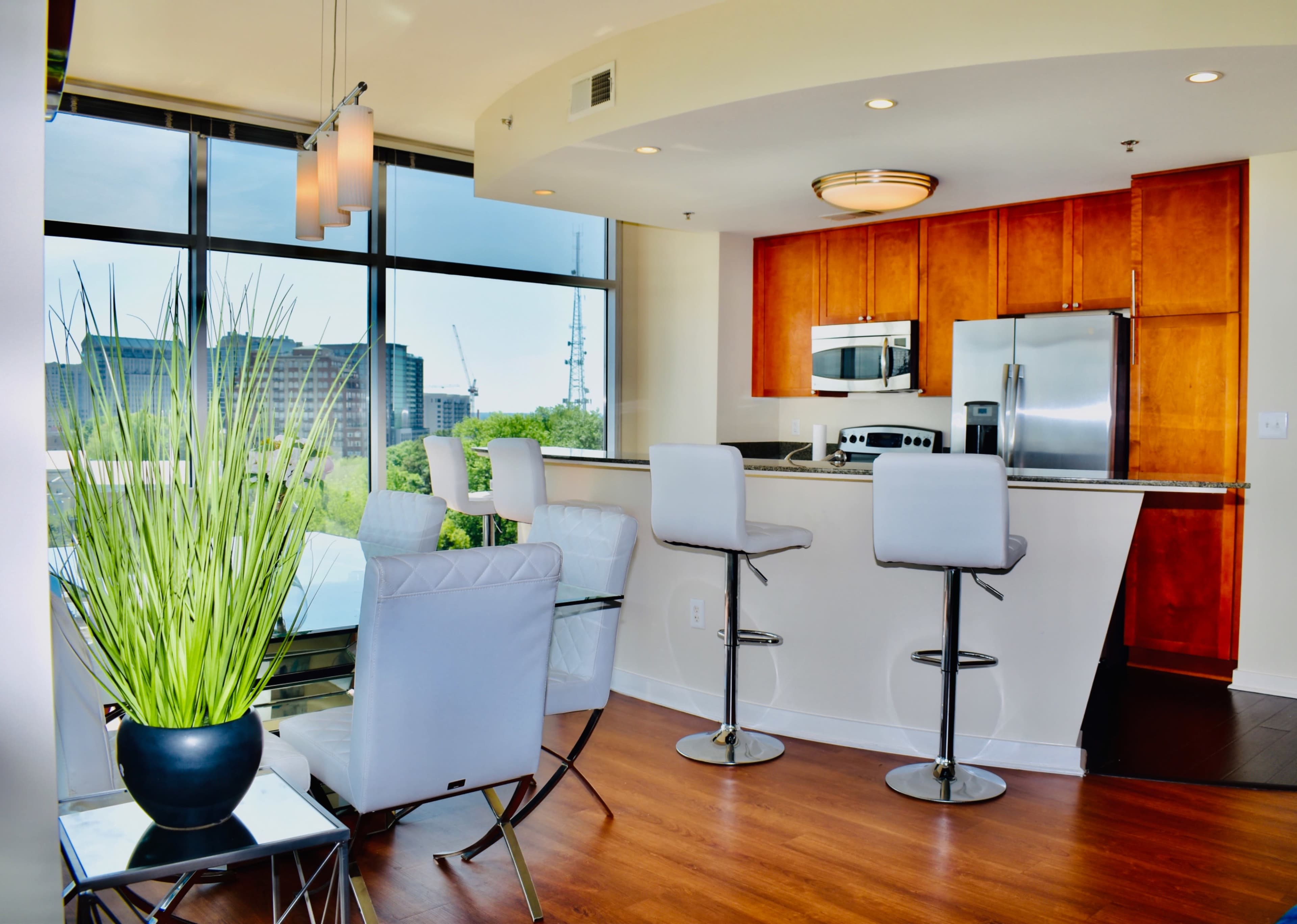 A modern kitchen features a bar with high stools, a dining area with a glass table, and large windows providing a view of the city.