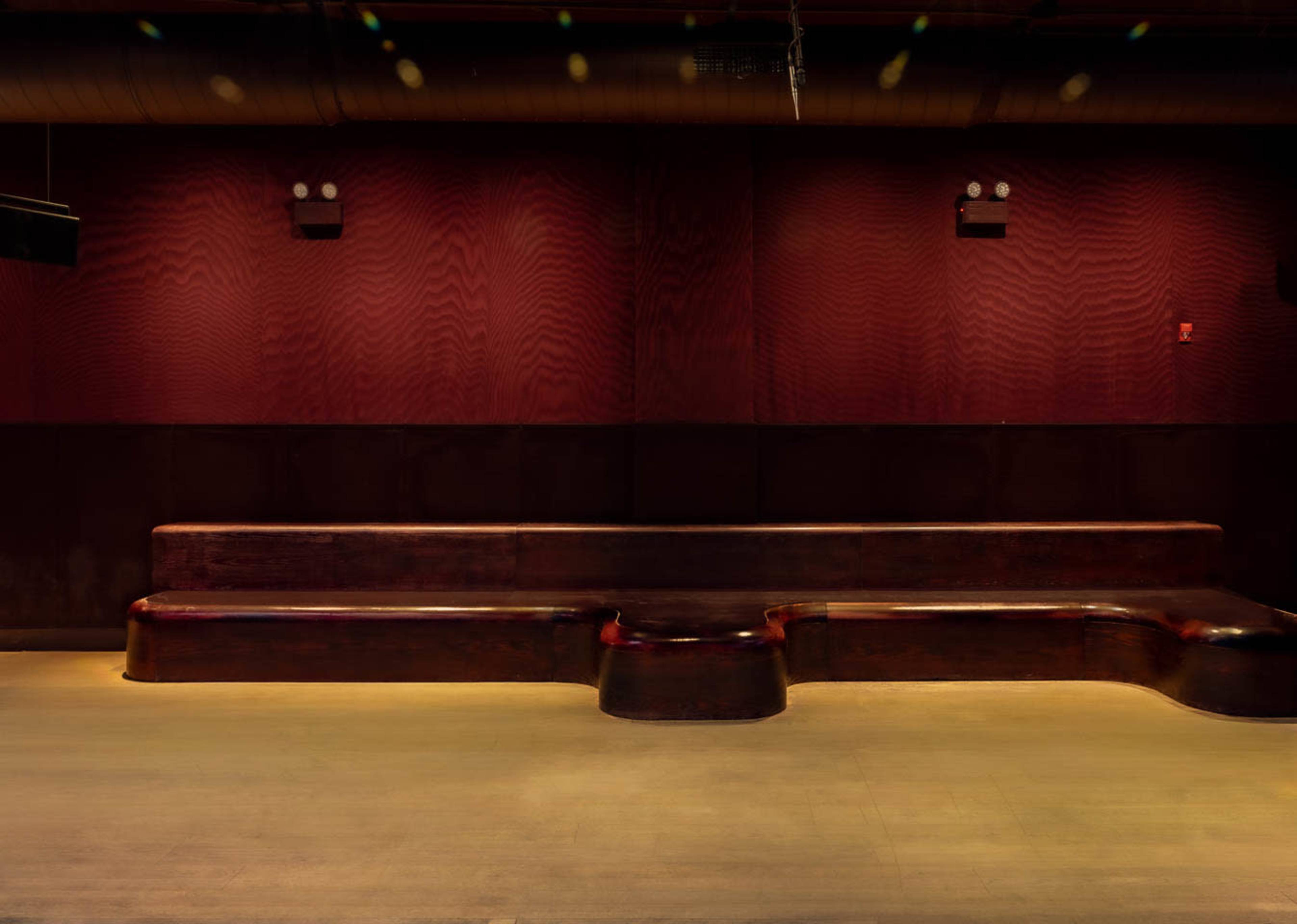 A long, dark wooden bench is positioned against a red wall in a dimly lit room.