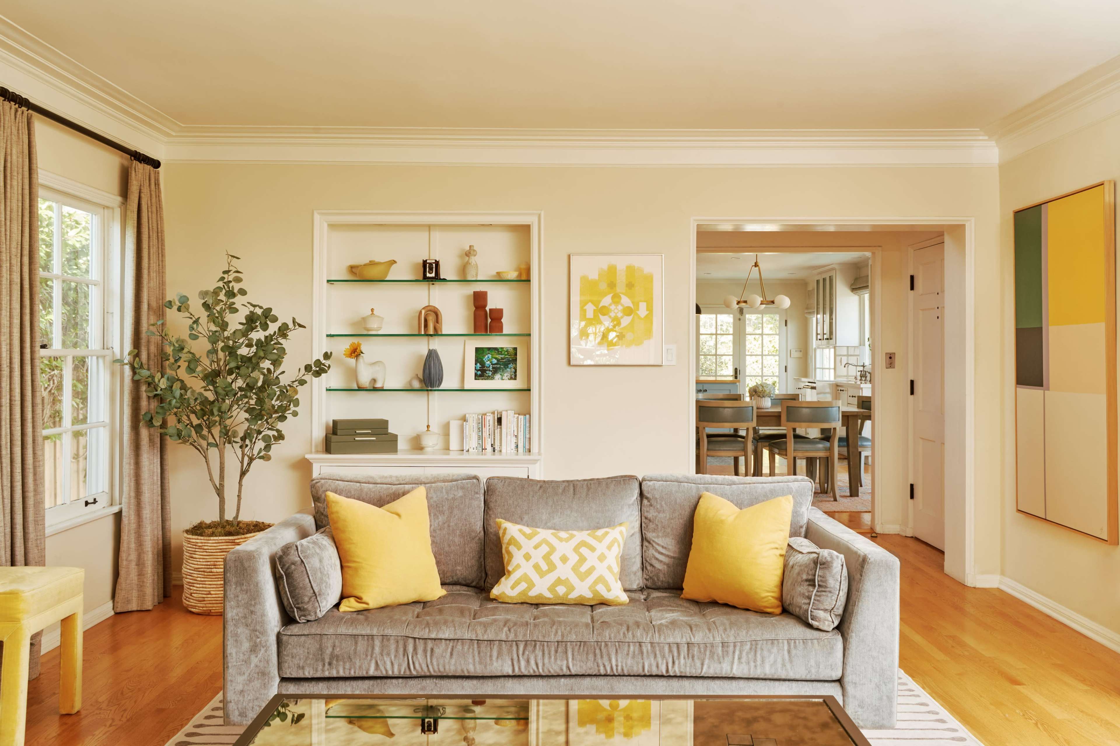 A gray sofa with yellow cushions is positioned in a well-lit living room featuring built-in shelves and a view of a dining area beyond.
