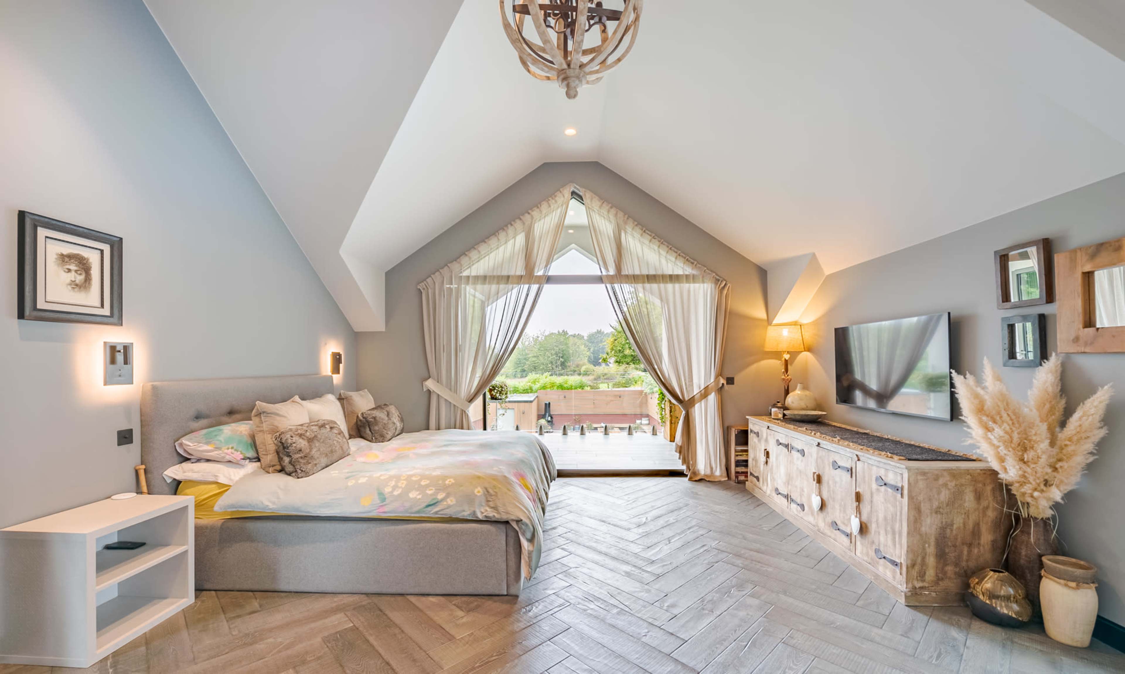 The bedroom features a large bed with decorative pillows, a wooden dresser, and a view of the outdoors through a large window.